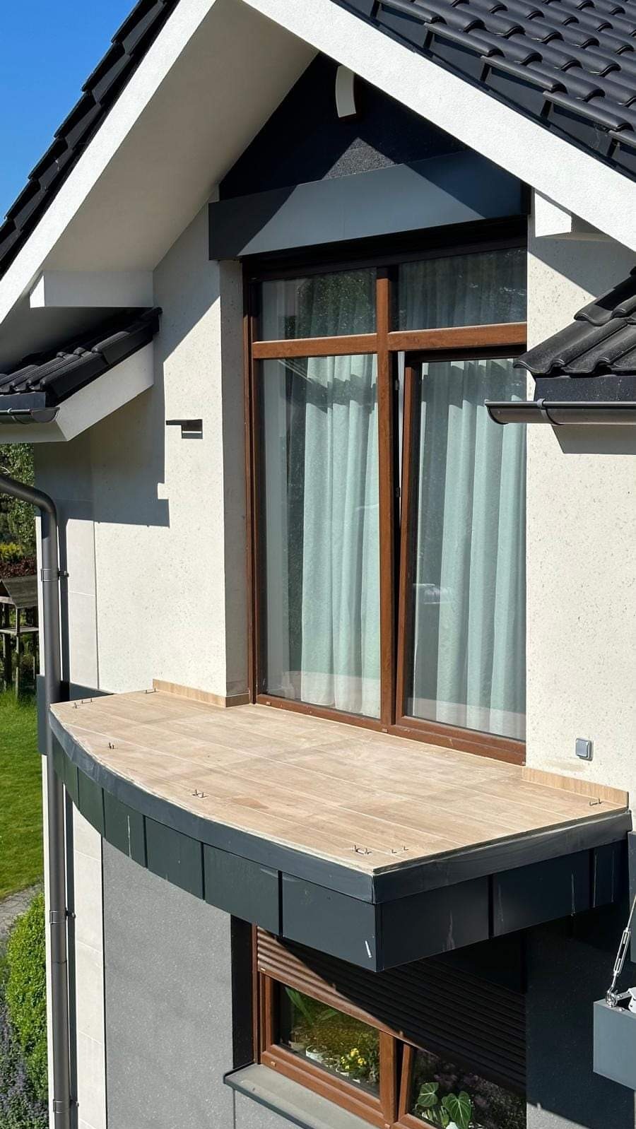 Nowoczesny balkon z drewnopodobną okładziną, czarne obramowanie, duże okno z brązowymi ramami i zasłonami, elewacja w jasnym odcieniu.