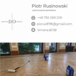 Parkiet ułożony w jodełkę po renowacji, widoczne dwa niebieskie pojemniki z preparatem i wałek malarski, wysokie okna z roślinami w tle.