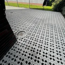 Szara ażurowa nawierzchnia z betonowych elementów, widoczny okrągły właz studzienki, w tle metalowe ogrodzenie i zieleń.
