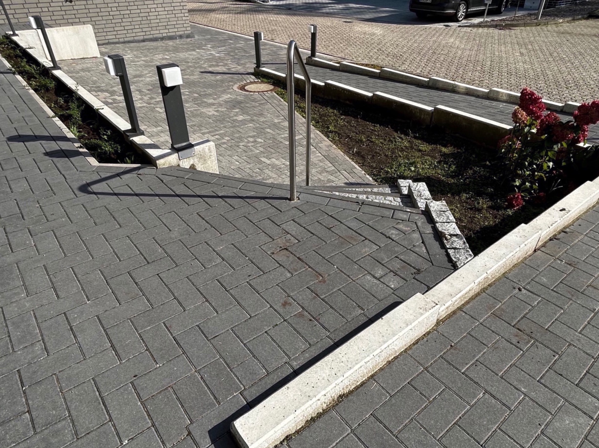 Ułożona szara kostka brukowa na schodach z metalową barierką, obok rabaty z roślinami i lampami oświetleniowymi, w tle chodnik z kostki i zaparkowany samochód.