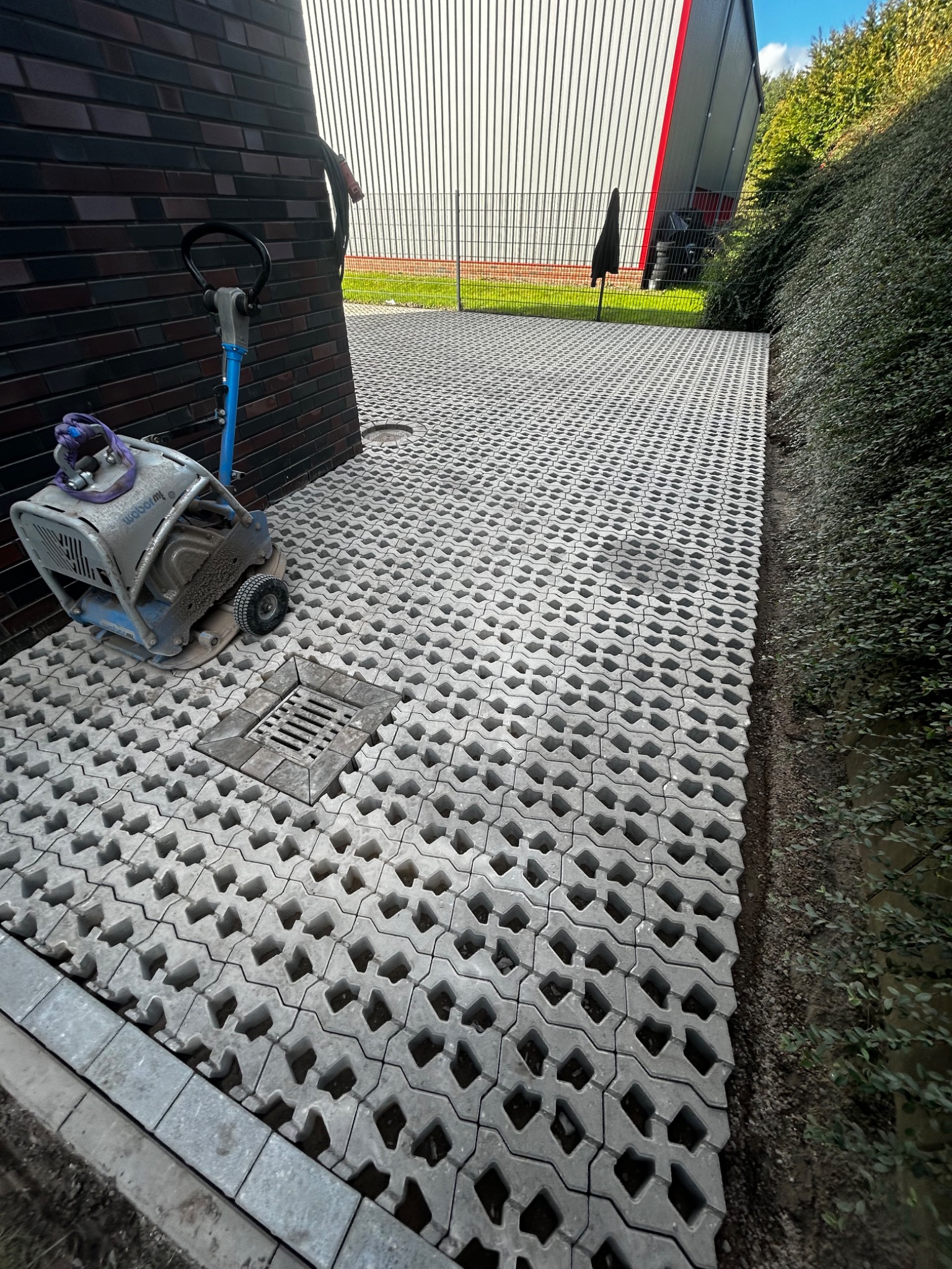 Ułożona szara ażurowa kostka brukowa na podjeździe, obok leży ubijarka wibracyjna i studzienka kanalizacyjna, w tle budynek z blachy trapezowej i żywopłot.
