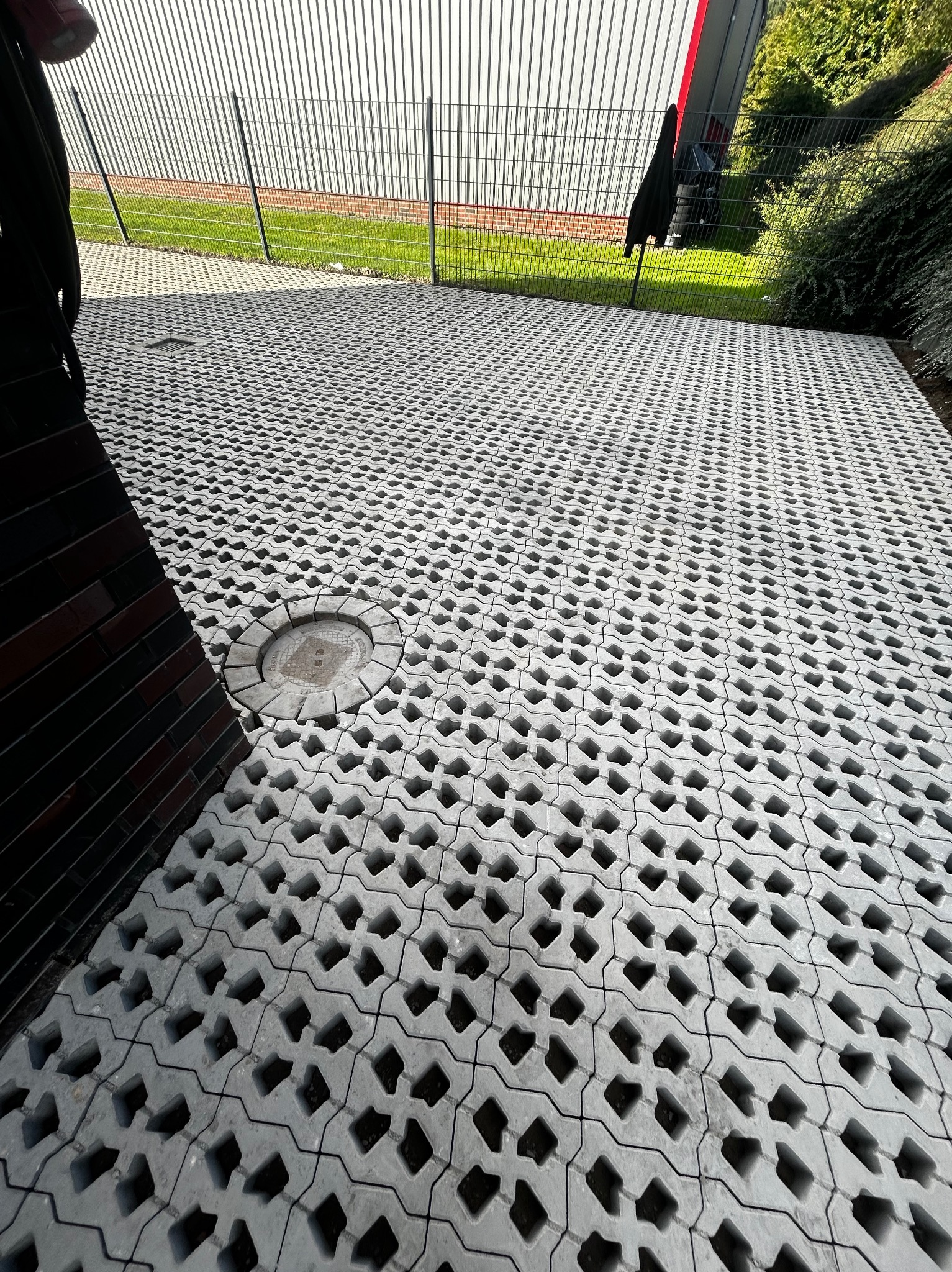 Szara ażurowa nawierzchnia z betonowych elementów, widoczny okrągły właz studzienki, w tle metalowe ogrodzenie i zieleń.