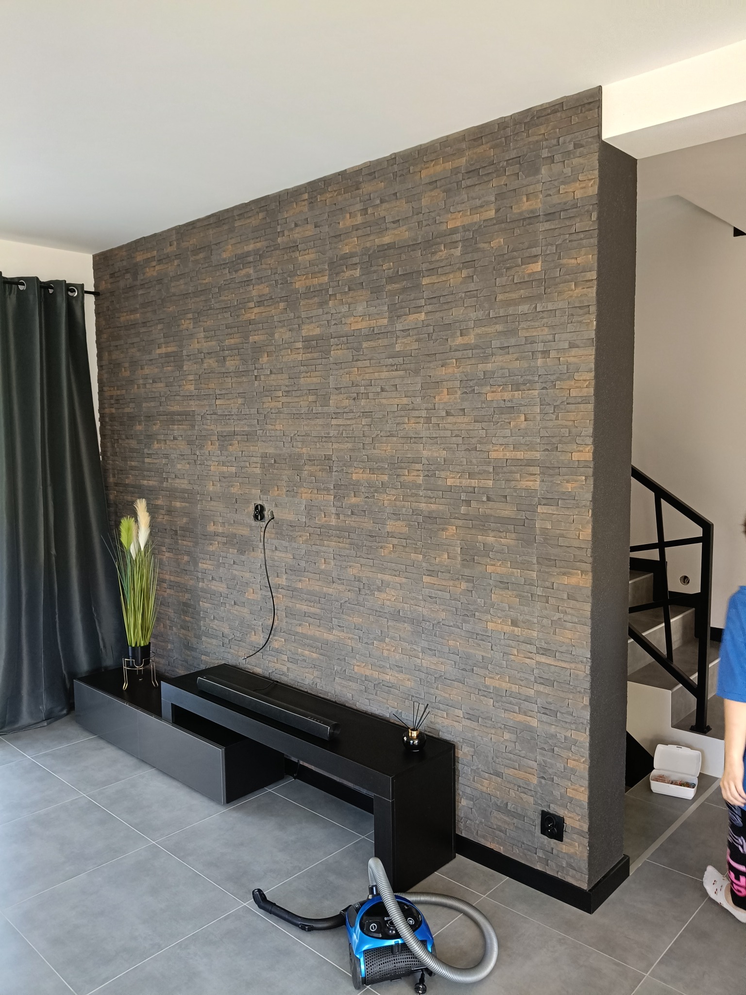 Ściana wykończona dekoracyjną okładziną imitującą kamień, czarny stolik RTV, szare płytki podłogowe, odkurzacz oraz fragment schodów w nowoczesnym wnętrzu.