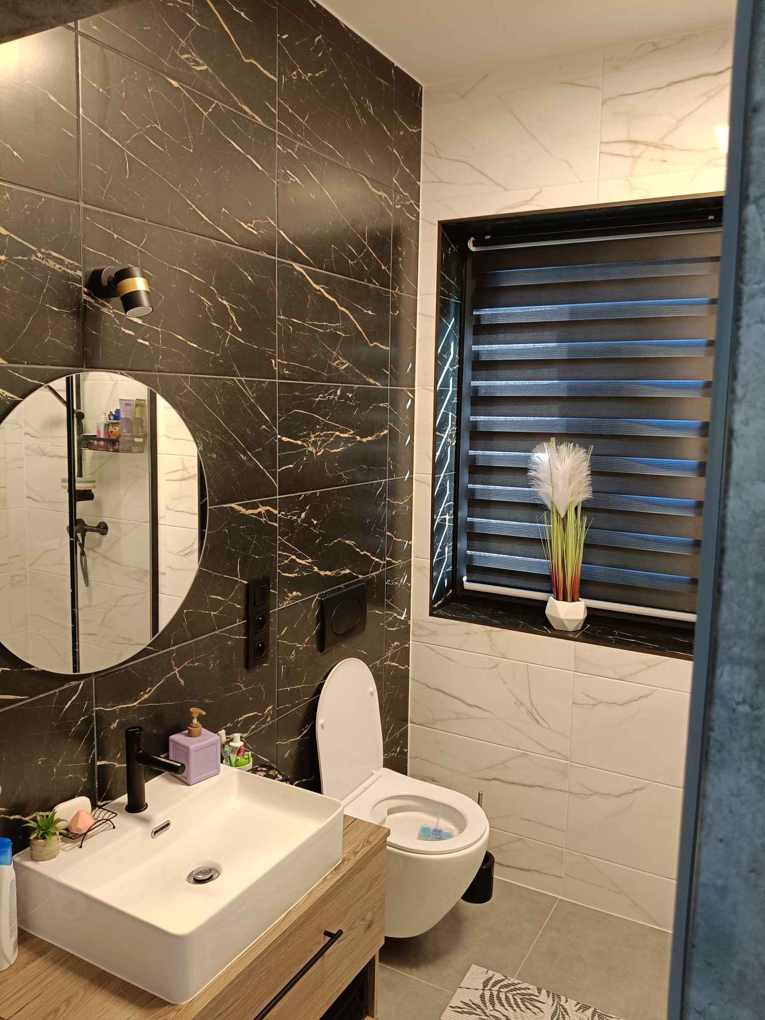 Nowoczesna łazienka z czarnymi płytkami imitującymi marmur na ścianie, okrągłym lustrem, białą umywalką nablatową na drewnianej szafce oraz oknem z żaluzjami dzień-noc.
