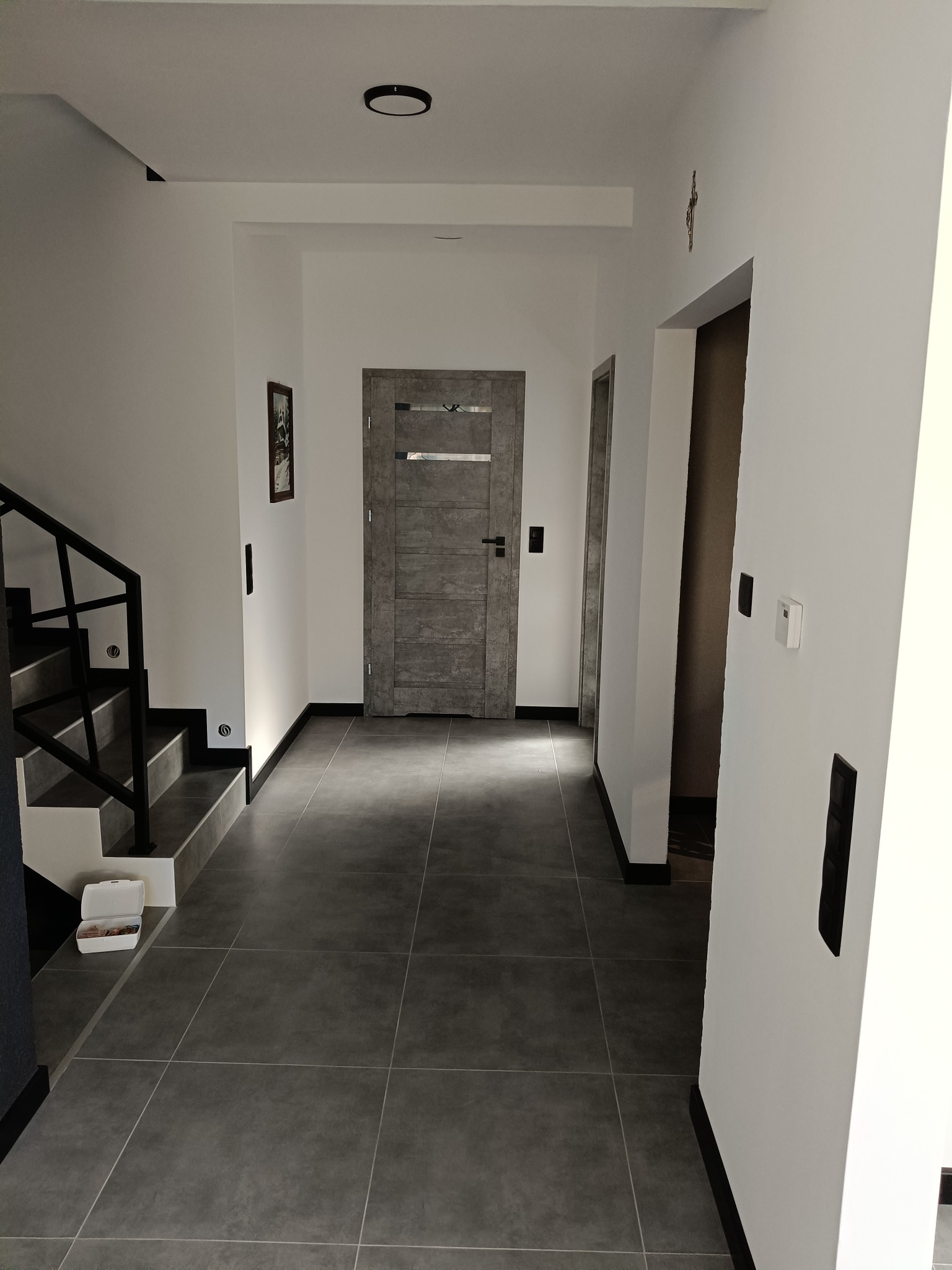 Wnętrze korytarza z szarymi płytkami podłogowymi, białe ściany z czarnymi listwami przypodłogowymi, schody z czarną balustradą, szare drzwi z poziomymi panelami i czarną klamką.