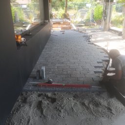 Ogrody Wojtala - Układanie szarej kostki brukowej wewnątrz budynku, widoczny fragment ułożonej nawierzchni, poziomica i pracownik w trakcie pracy. W tle widoczne materiały budowlane.
