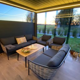 Ogrody Wojtala - Zadaszony taras z meblami ogrodowymi z rattanu i drewnianym stołem. Widok na ogród i zachodzące słońce przez szklane ściany. Oświetlenie LED pod dachem.