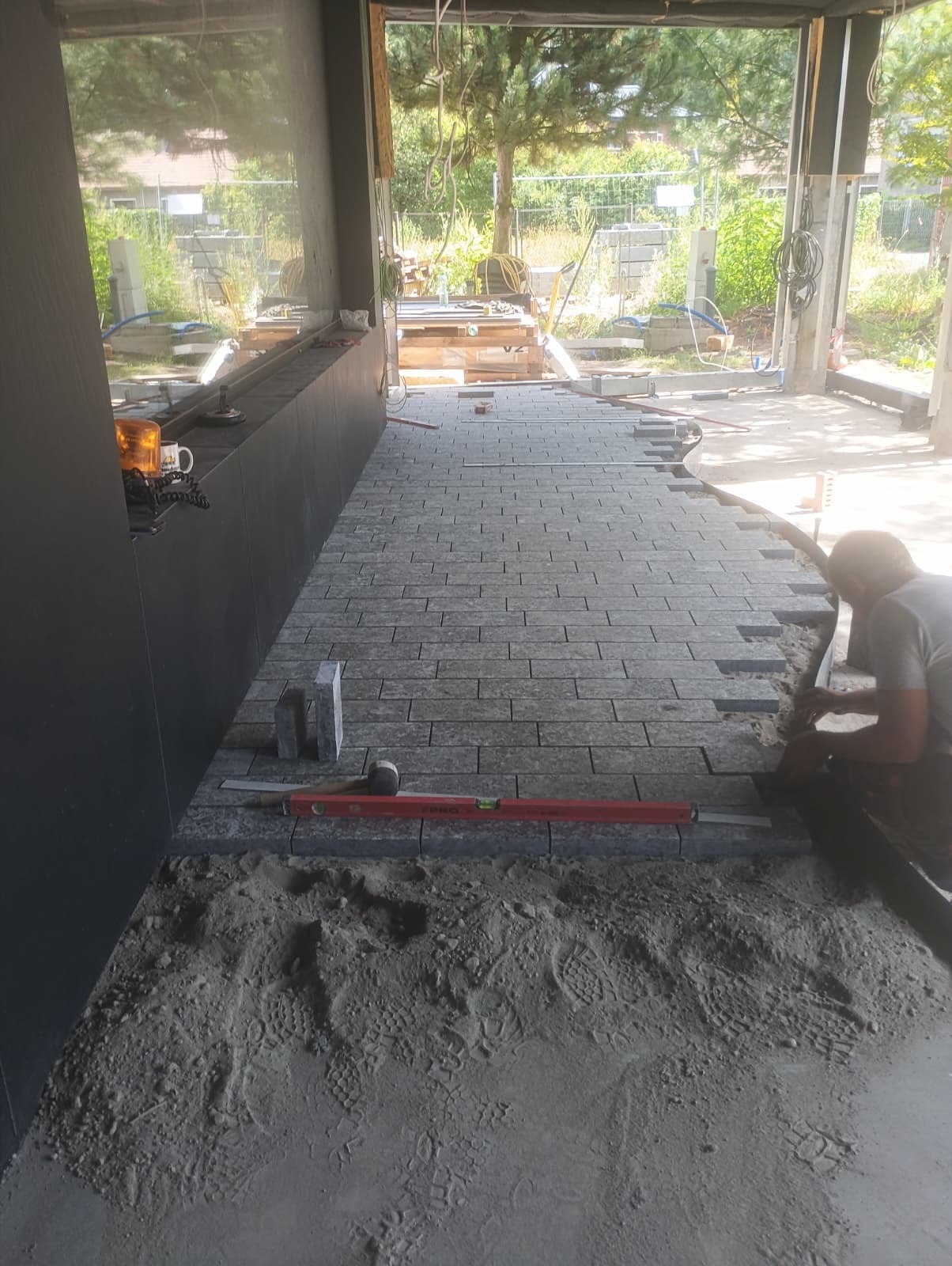 Układanie szarej kostki brukowej wewnątrz budynku, widoczny fragment ułożonej nawierzchni, poziomica i pracownik w trakcie pracy. W tle widoczne materiały budowlane.