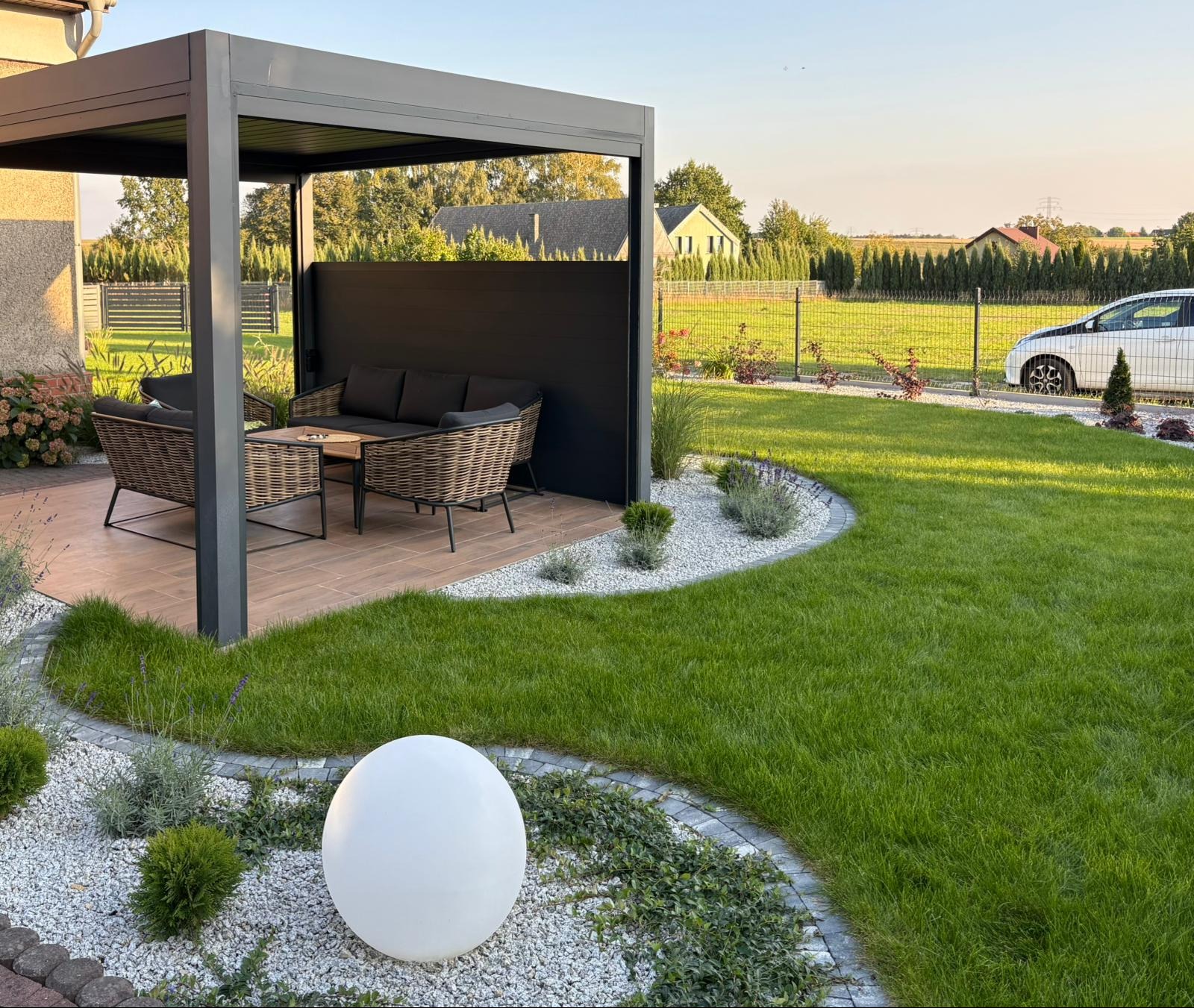 Nowoczesny ogród z pergolą, meblami ogrodowymi i oświetleniem kulistym. Starannie utrzymany trawnik, obrzeża z kamienia i rośliny ozdobne tworzą harmonijną całość.