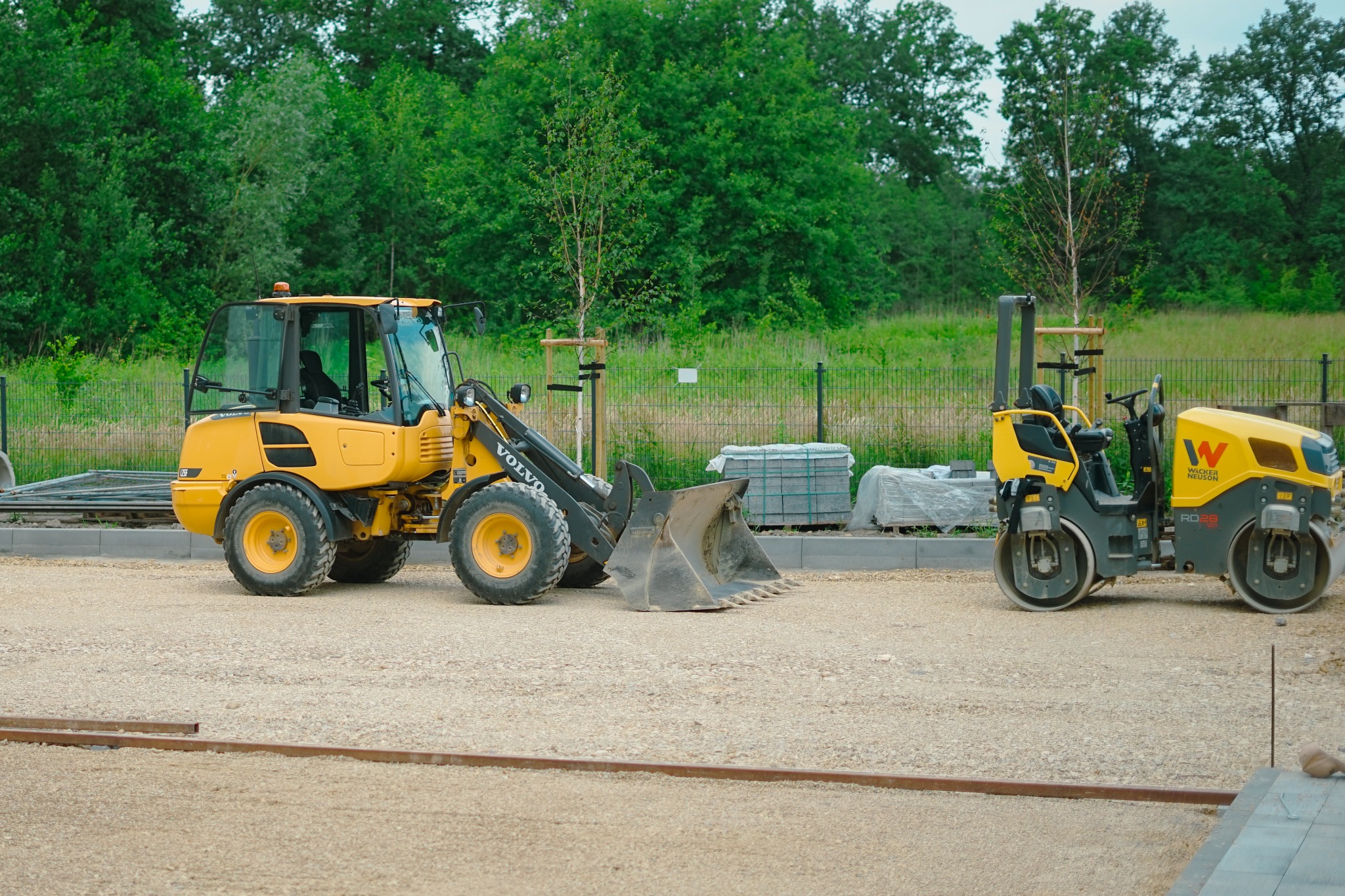 Żółta ładowarka Volvo i walec Wacker Neuson na żwirowym podłożu, w tle zieleń i kostka brukowa. Przygotowanie terenu pod budowę.