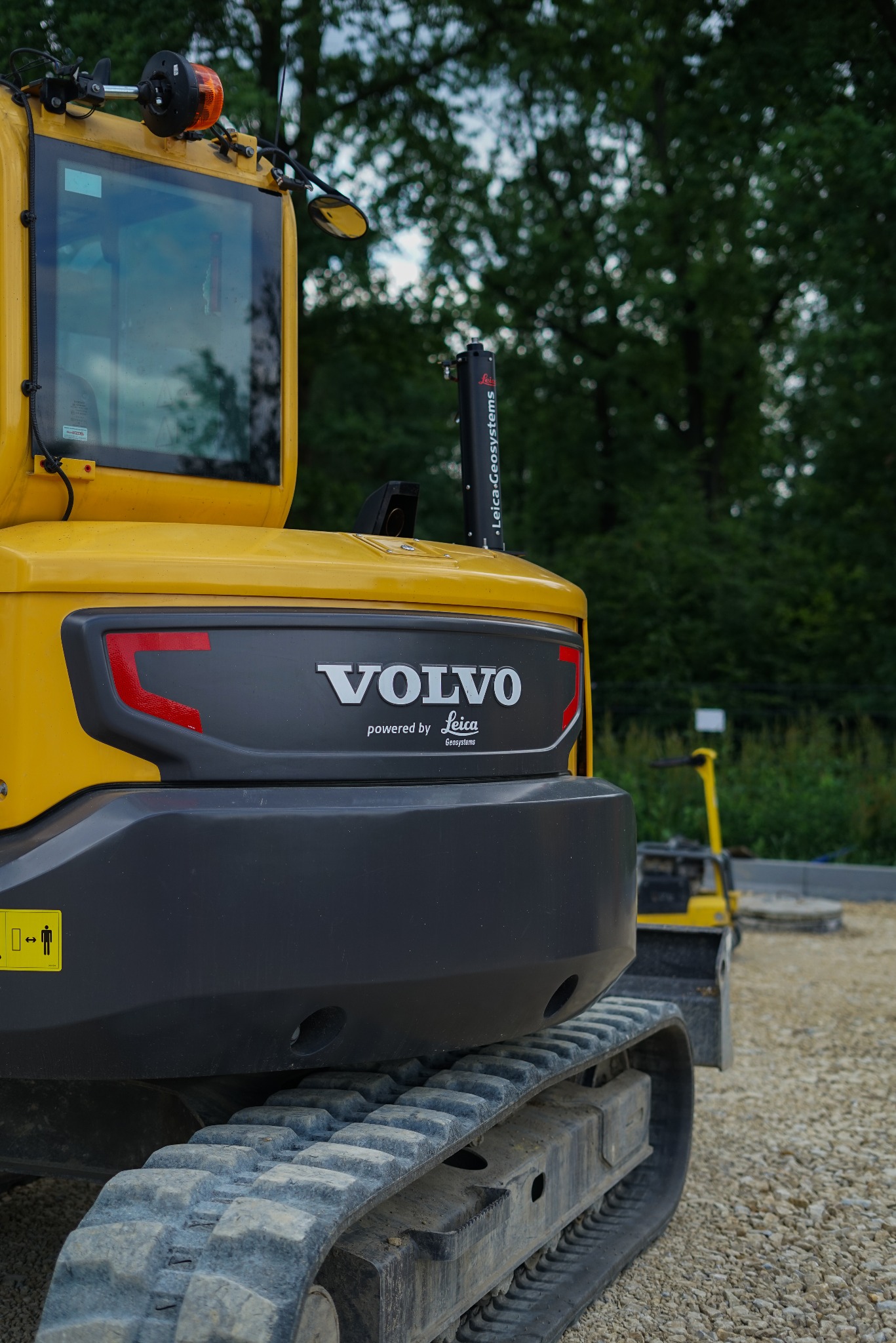 Żółta koparka Volvo z systemem Leica Geosystems na żwirowym podłożu, w tle zieleń i ubijarka. Widoczny fragment gąsienicy i tabliczka informacyjna.