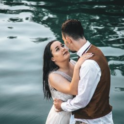 Zatrzymać Chwile - Para młoda w objęciach na tle wody, ona w białej sukni z kryształkami, on w brązowej kamizelce, woda odbija niebo.