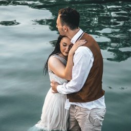 Zatrzymać Chwile - Para młoda, ubrana w suknię ślubną i brązową kamizelkę, stoi objęta w wodzie jeziora, z odbiciami światła na powierzchni wody.