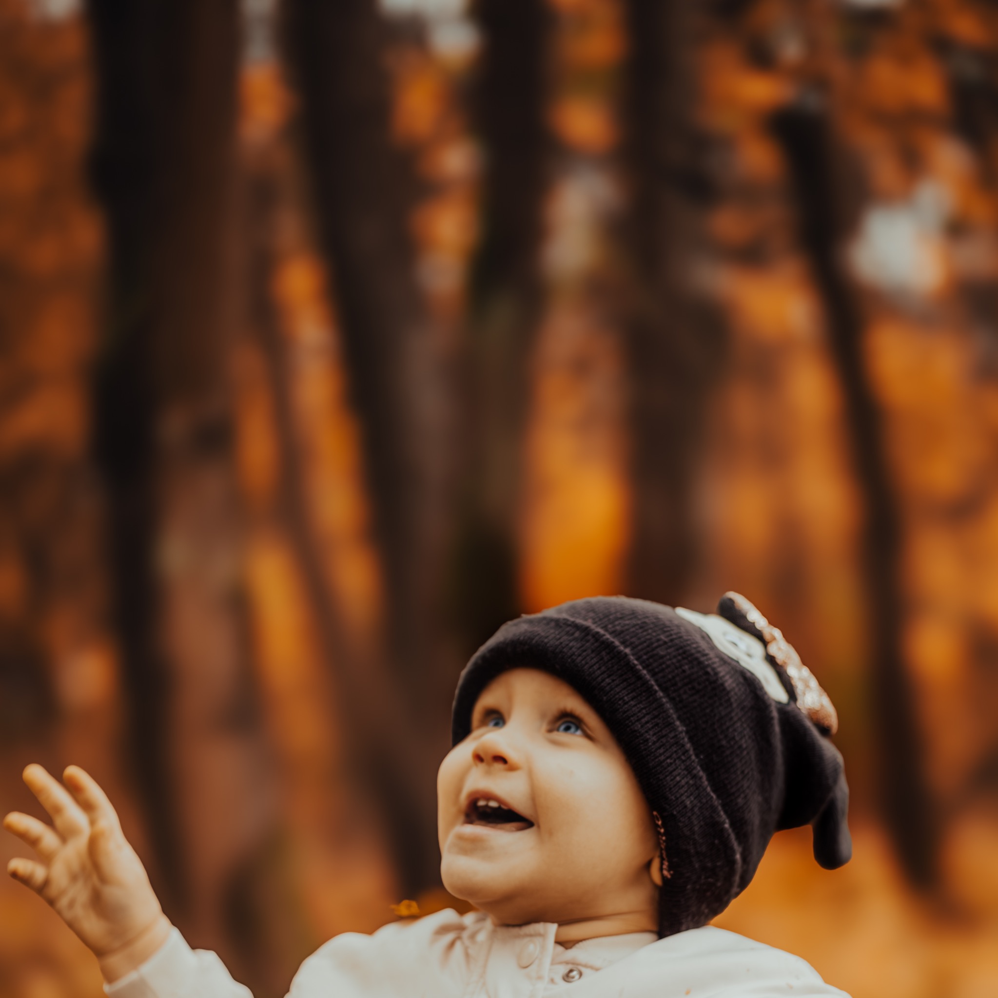 Uśmiechnięta dziewczynka w czapce z błyszczącą aplikacją, spoglądająca w górę na tle jesiennego lasu z rozmazanymi drzewami i opadającymi liśćmi.