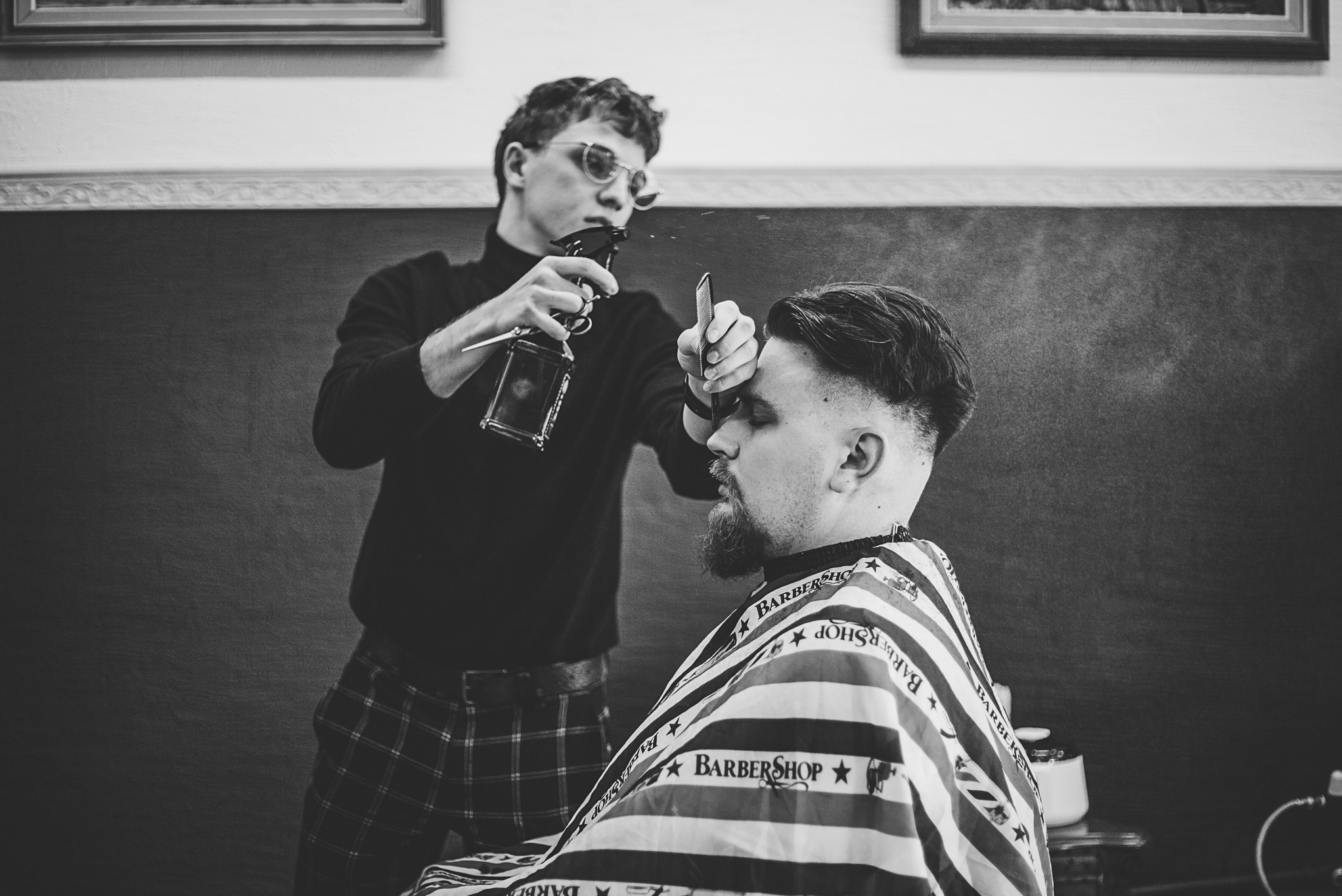 Czarno-białe zdjęcie fryzjera w okularach, spryskującego włosy klienta przed strzyżeniem. Klient siedzi w fotelu, okryty peleryną w paski z napisem 'Barbershop'.