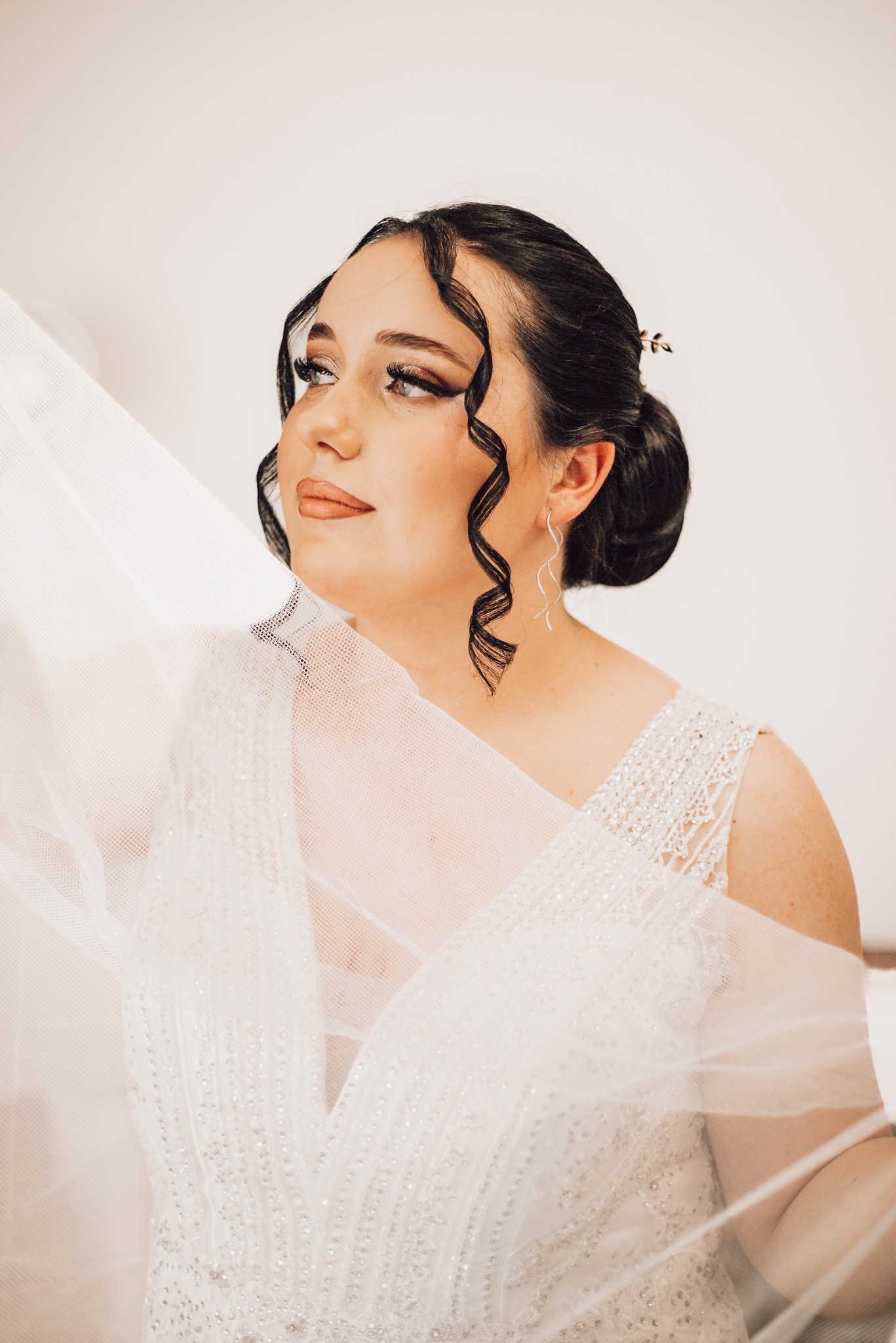 Panna młoda w sukni ślubnej z welonem, upięte ciemne włosy z ozdobną spinką, delikatny makijaż, srebrne kolczyki w kształcie fali, patrzy w bok.