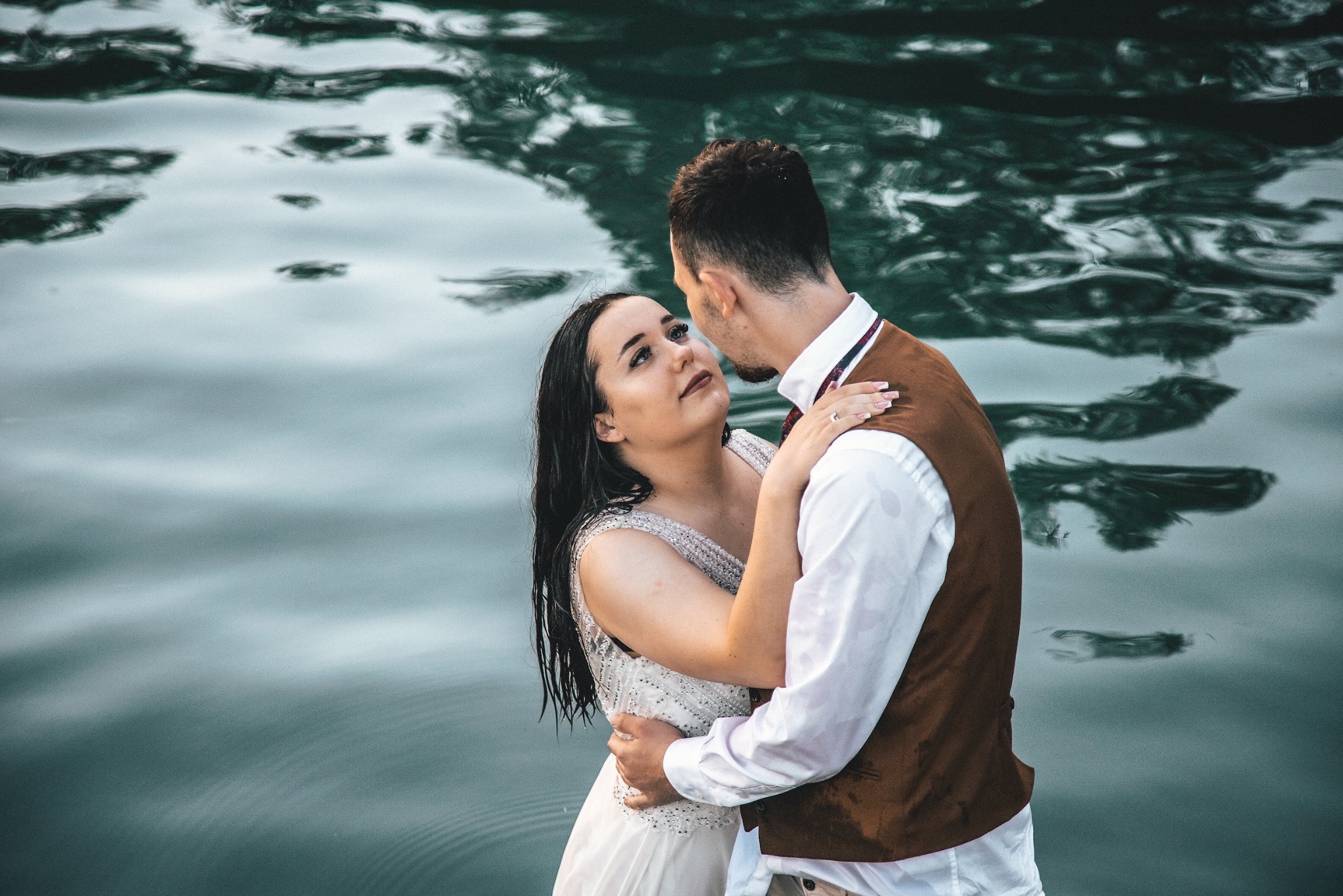 Para młoda w objęciach na tle wody, ona w białej sukni z kryształkami, on w brązowej kamizelce, woda odbija niebo.