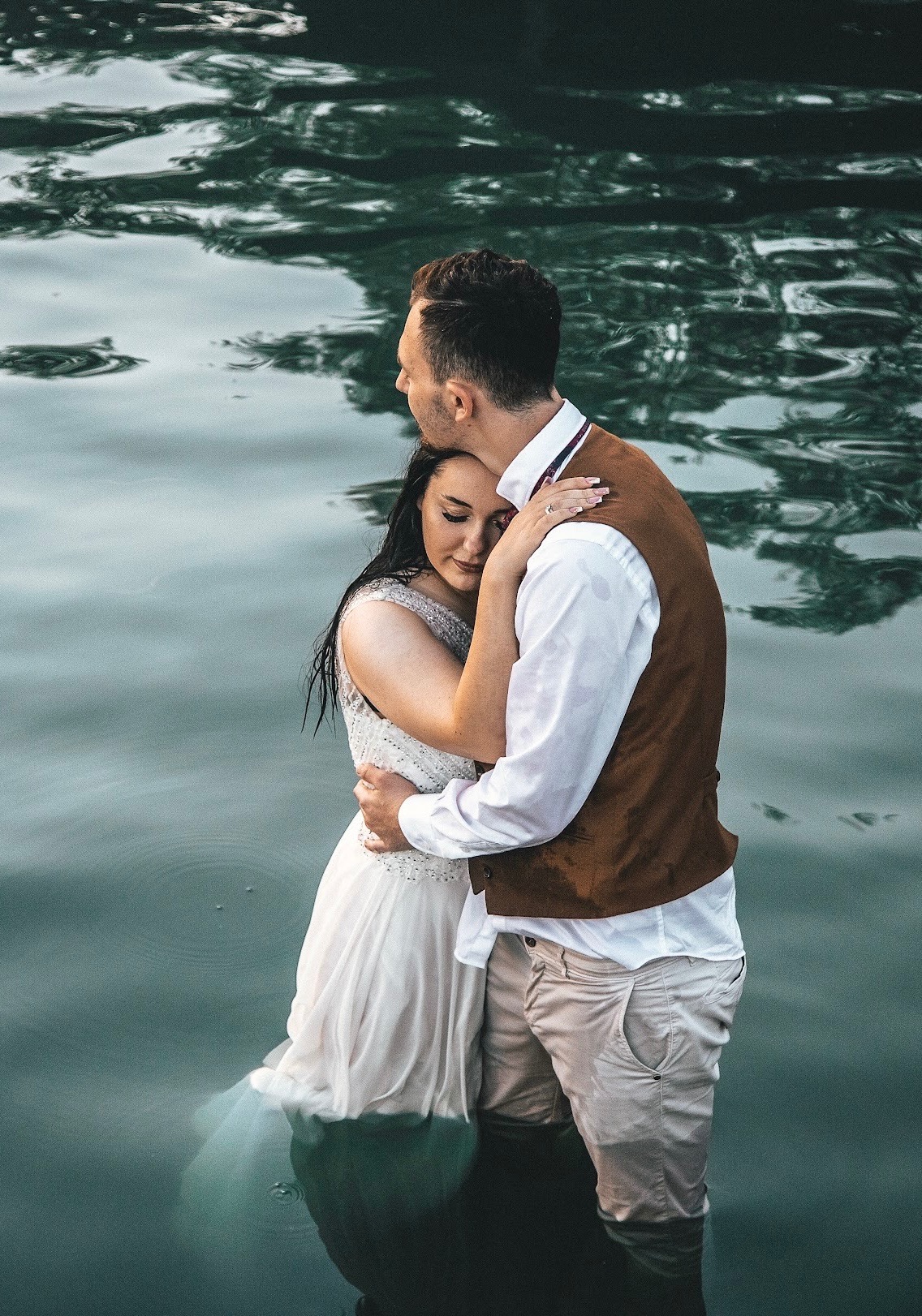 Para młoda, ubrana w suknię ślubną i brązową kamizelkę, stoi objęta w wodzie jeziora, z odbiciami światła na powierzchni wody.
