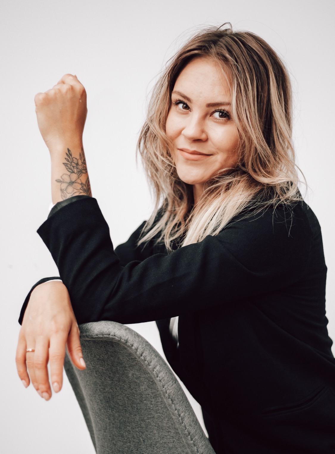 Portret uśmiechniętej kobiety w czarnej marynarce, opierającej się o szare krzesło, z widocznym tatuażem z motywem kwiatowym na przedramieniu.