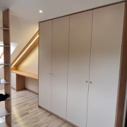 Wnękowa szafa z czterema drzwiami w kolorze beżowym, z drewnianym obramowaniem i okrągłymi uchwytami, zintegrowana z biurkiem pod skosem dachu, oświetlonym taśmą LED, na podłodze panele imitujące...