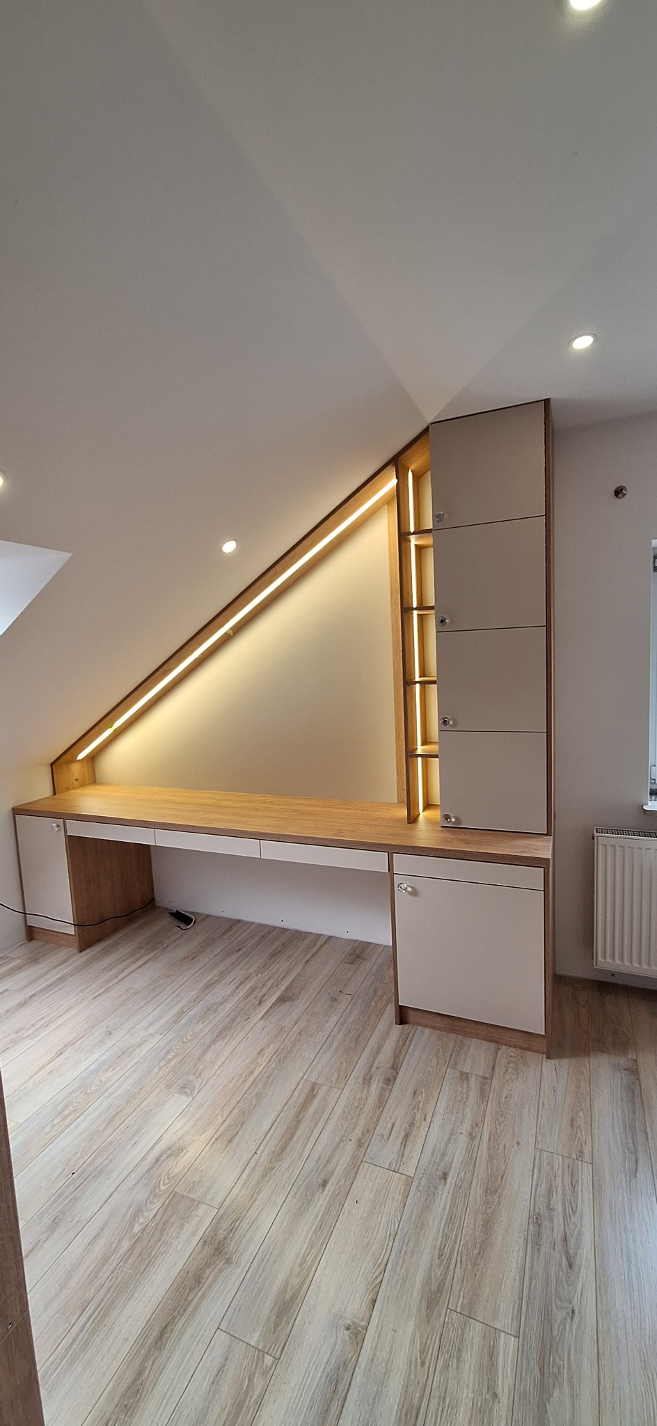 Biurko na wymiar wkomponowane w skos dachu, z oświetleniem LED i szafkami, jasne drewno i białe fronty, drewniana podłoga.