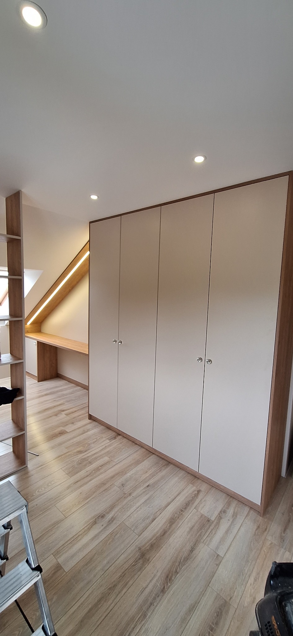 Wnękowa szafa z czterema drzwiami w kolorze beżowym, z drewnianym obramowaniem i okrągłymi uchwytami, zintegrowana z biurkiem pod skosem dachu, oświetlonym taśmą LED, na podłodze panele imitujące...