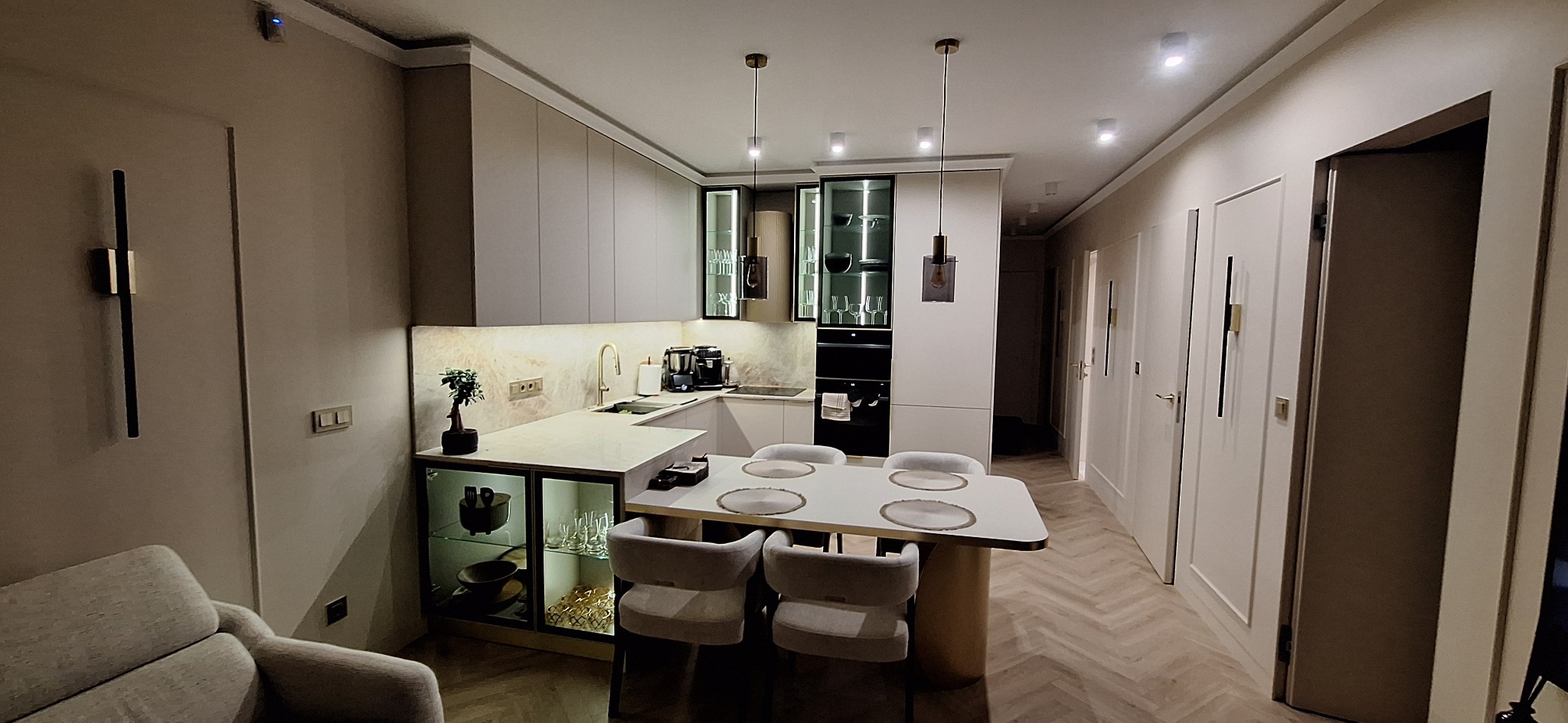 Nowoczesna kuchnia w odcieniach beżu z przeszklonymi szafkami, oświetleniem punktowym i stołem jadalnym. Parkiet w jodełkę. Minimalistyczny design.