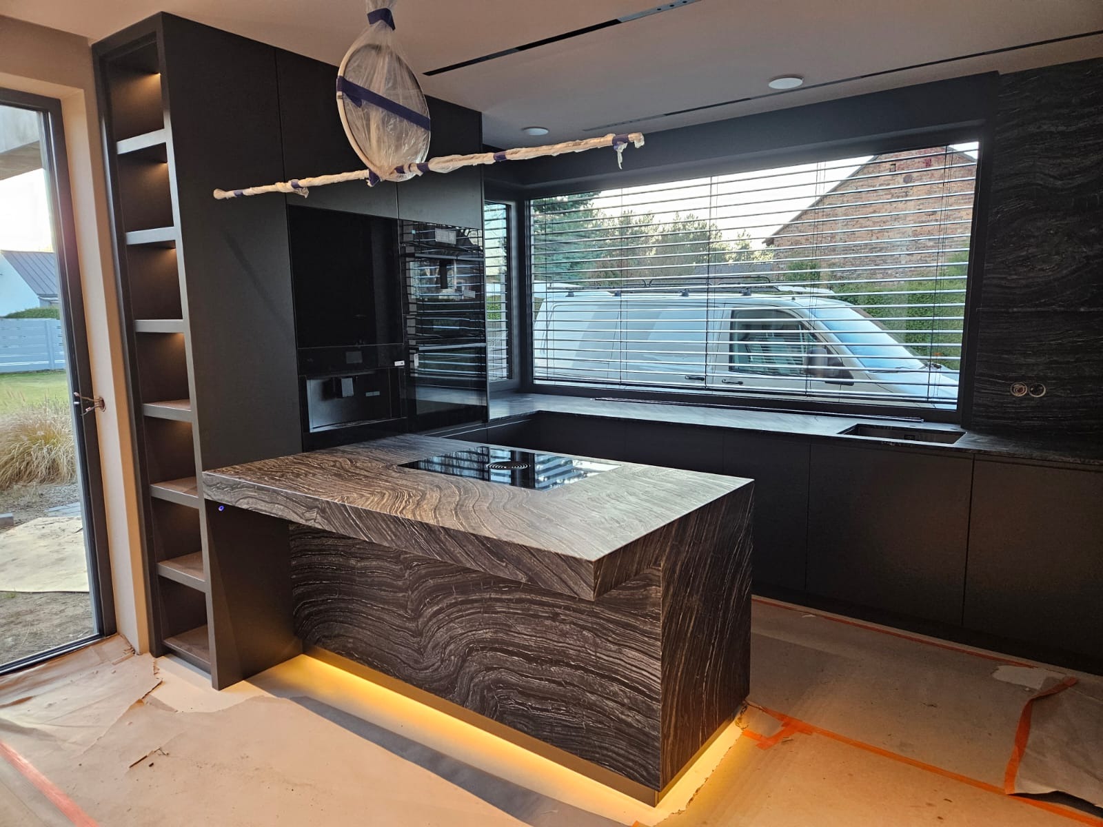 Nowoczesna, czarna kuchnia z wyspą o unikalnym wzorze kamienia i podświetleniem LED. Widok na zewnątrz przez okno z żaluzjami. Elementy w trakcie montażu.