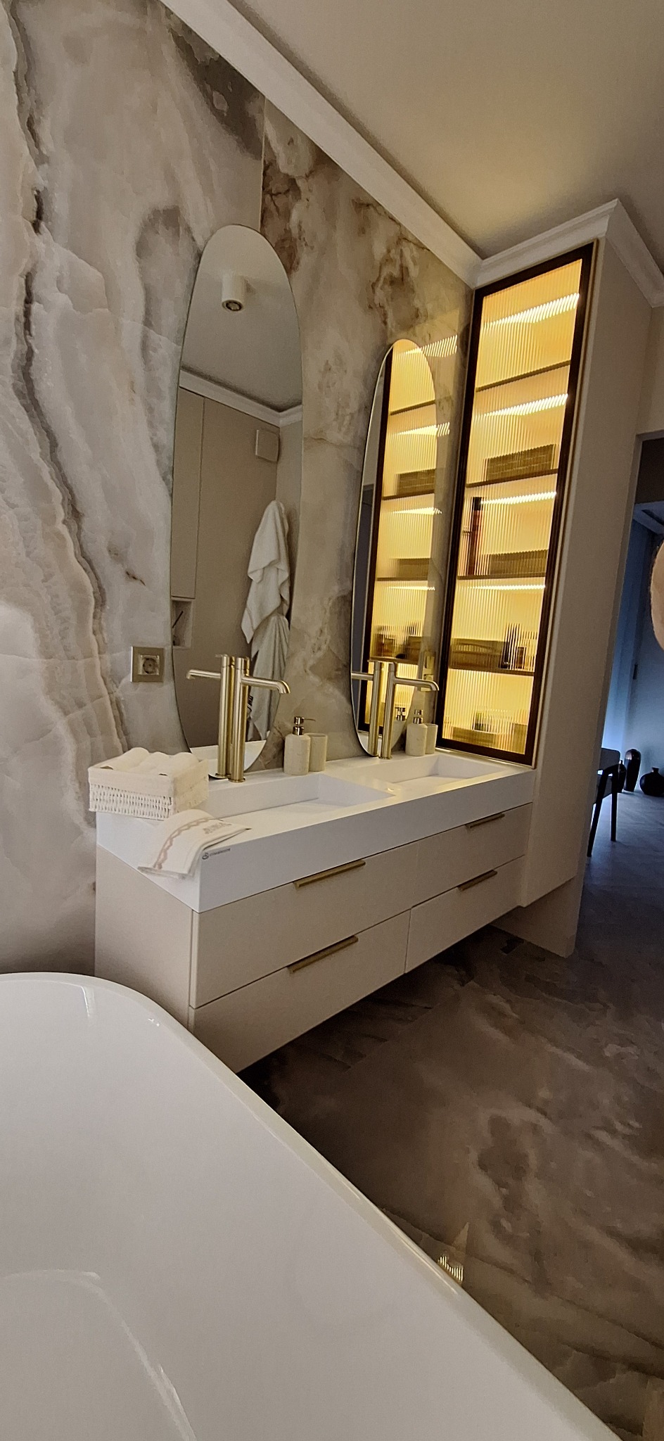 Elegancka łazienka z podwójną umywalką, złotymi bateriami i szafką z oświetleniem. Marmurowe ściany i podłoga dodają luksusowego charakteru.