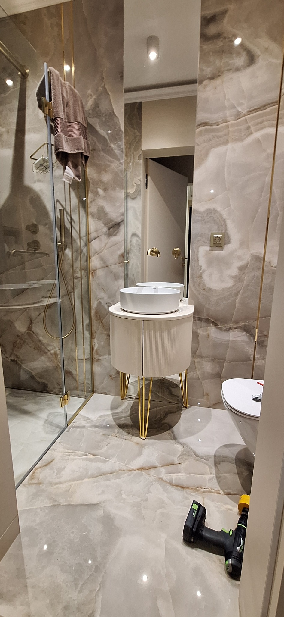 Nowoczesna łazienka z umywalką na szafce z ryflowanymi drzwiczkami i złotymi nóżkami. Na podłodze leży wkrętarka. Marmurowe ściany i złote akcenty.