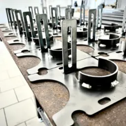 Metalowe elementy po cięciu i spawaniu, ułożone rzędami na stole warsztatowym, widoczne ślady spawania i obróbki.