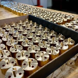 DYNAMET Daniel Barborka - Partie mosiężnych, cylindrycznych elementów z trzpieniem i otworami, ułożonych w kartonowym pudełku i na drewnianej powierzchni, widoczne ślady obróbki.