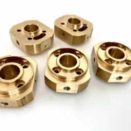 Pięć precyzyjnie wykonanych, złotych elementów metalowych z otworami, ułożonych na białym tle. Widoczne ślady obróbki mechanicznej i gwintowane otwory.