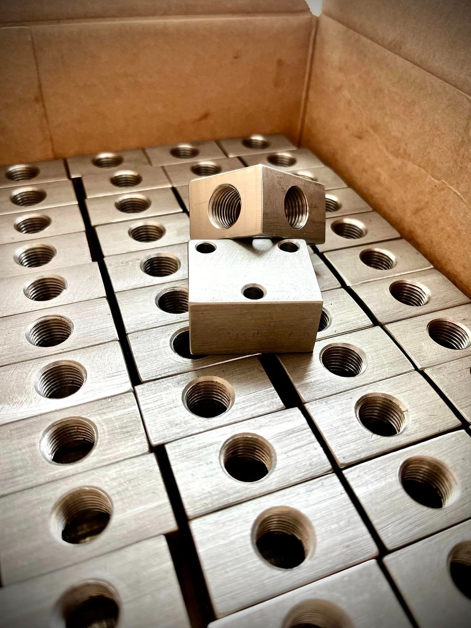 Partia obrobionych, metalowych bloków z gwintowanymi otworami ułożonych w kartonowym pudełku, dwa bloki ułożone jeden na drugim na wierzchu.