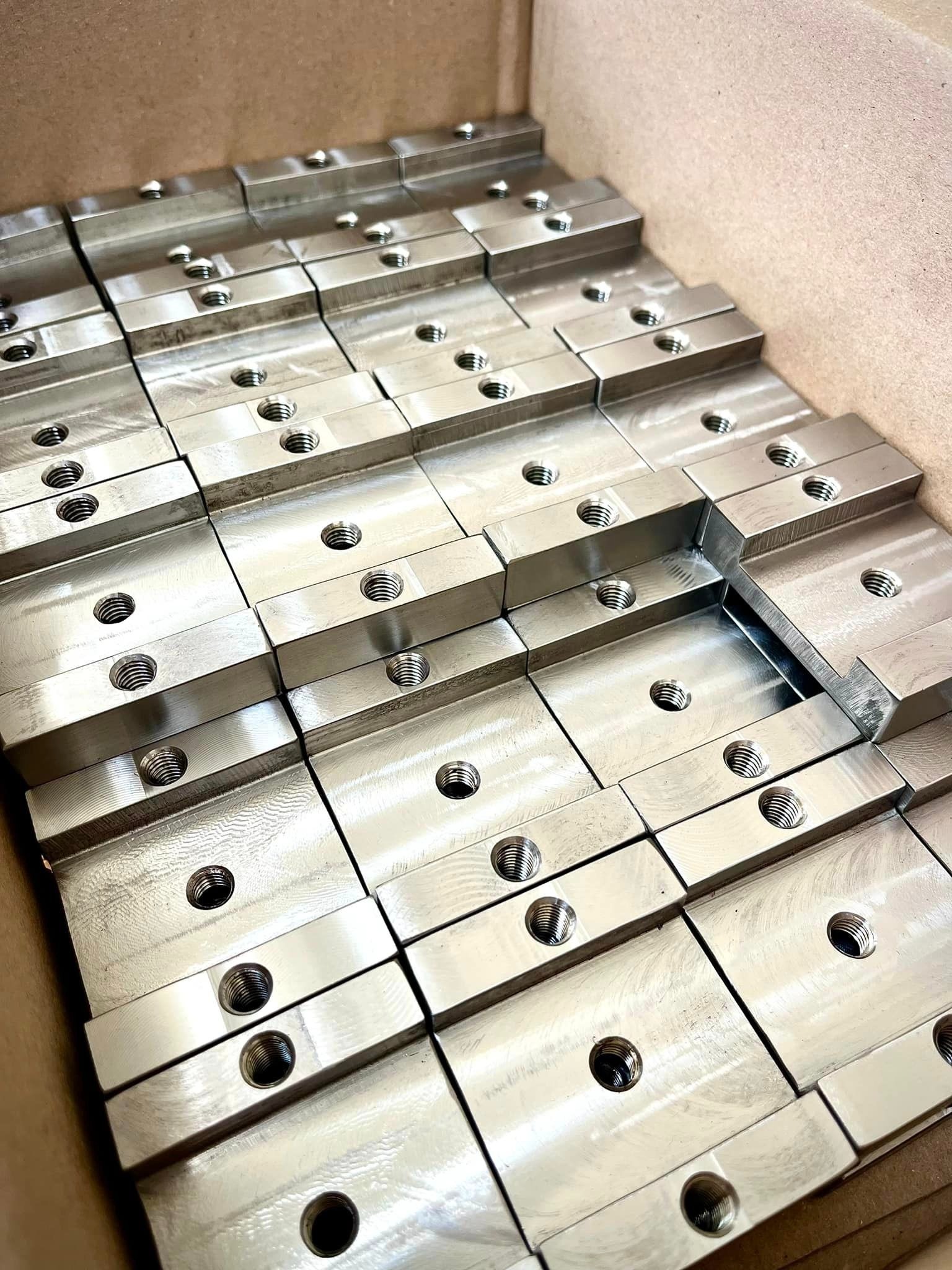 Partia precyzyjnie obrobionych, metalowych elementów z gwintowanymi otworami, ułożonych warstwami w kartonowym pudełku, widoczne ślady obróbki na powierzchni.