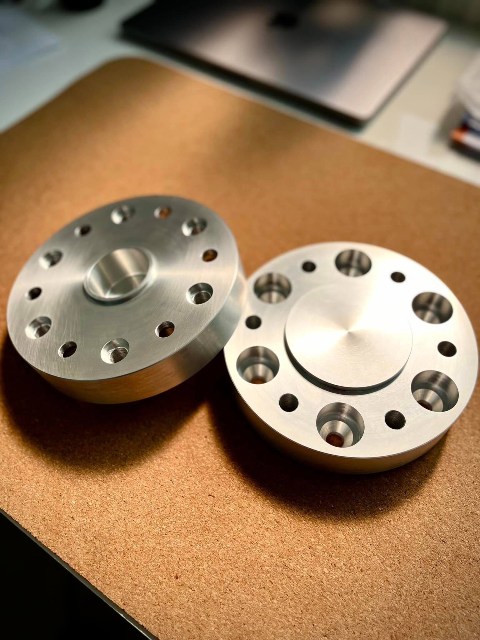 Dwa precyzyjnie wykonane, srebrne, okrągłe elementy z aluminium z otworami, leżące na brązowej powierzchni biurka, widoczne ślady obróbki na powierzchni.