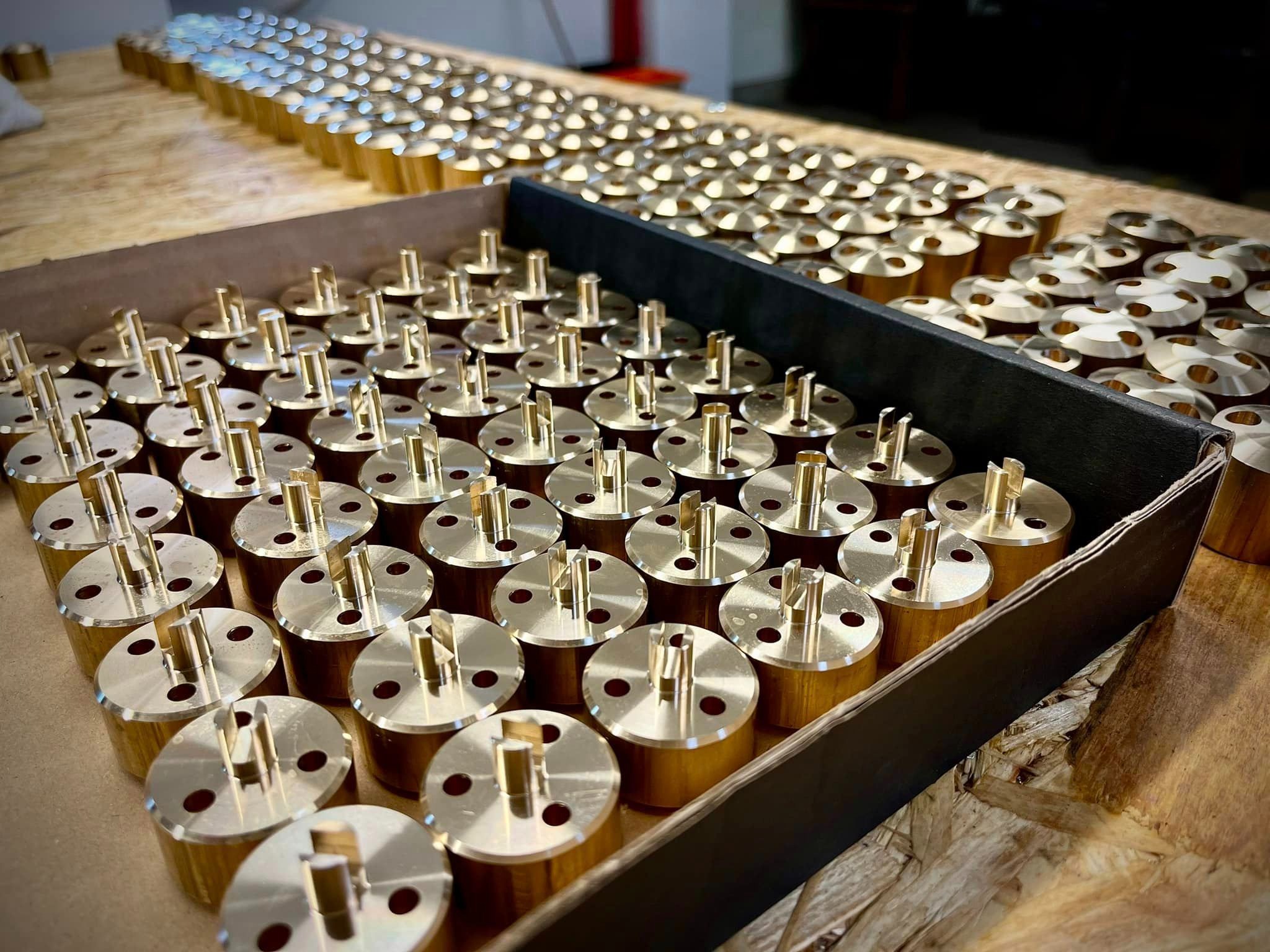 Partie mosiężnych, cylindrycznych elementów z trzpieniem i otworami, ułożonych w kartonowym pudełku i na drewnianej powierzchni, widoczne ślady obróbki.