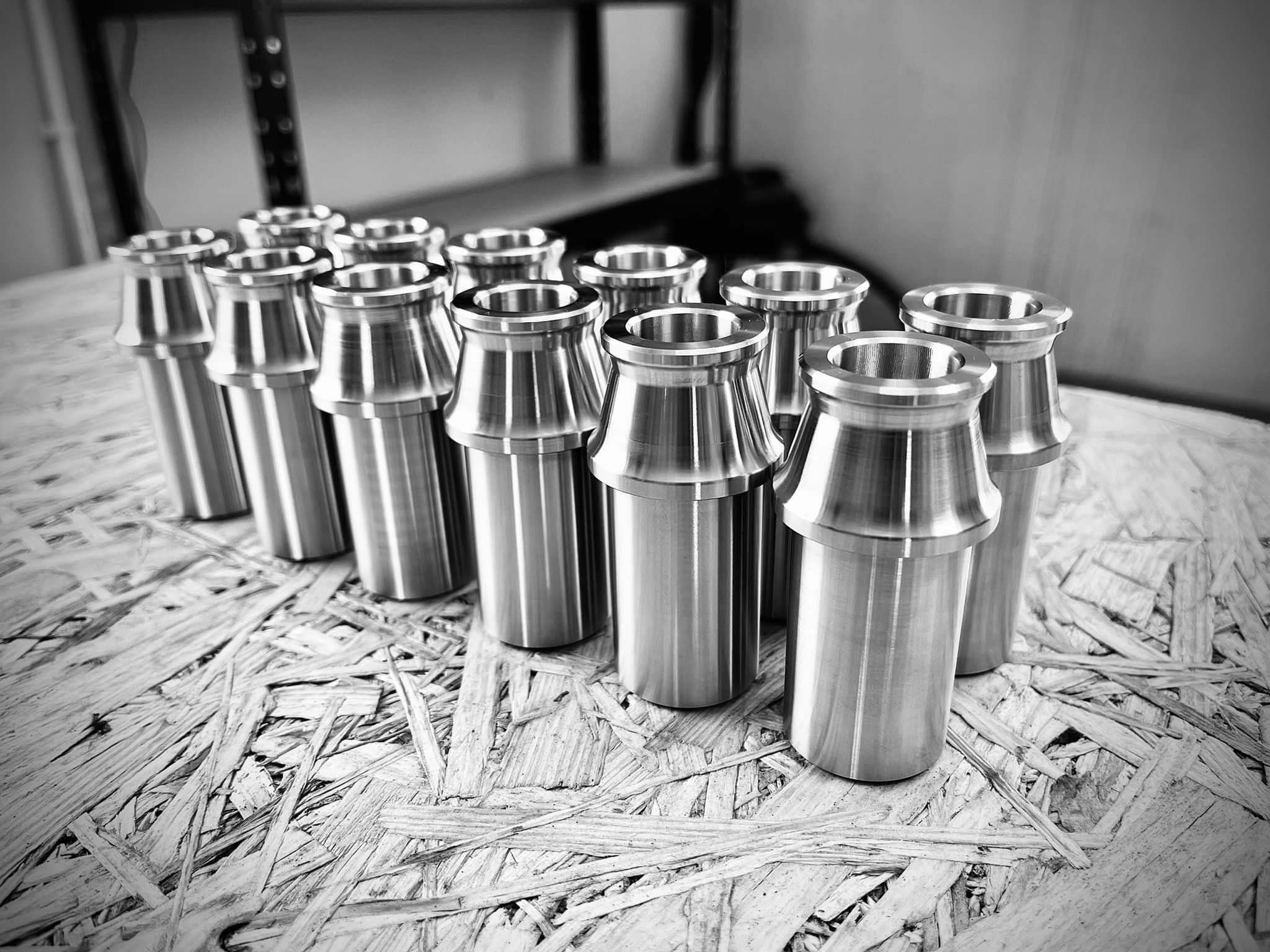 Czarno-białe zdjęcie kilkunastu identycznych, metalowych elementów z kołnierzem, ustawionych w rzędach na płycie OSB. Elementy są gładkie i mają cylindryczny kształt.