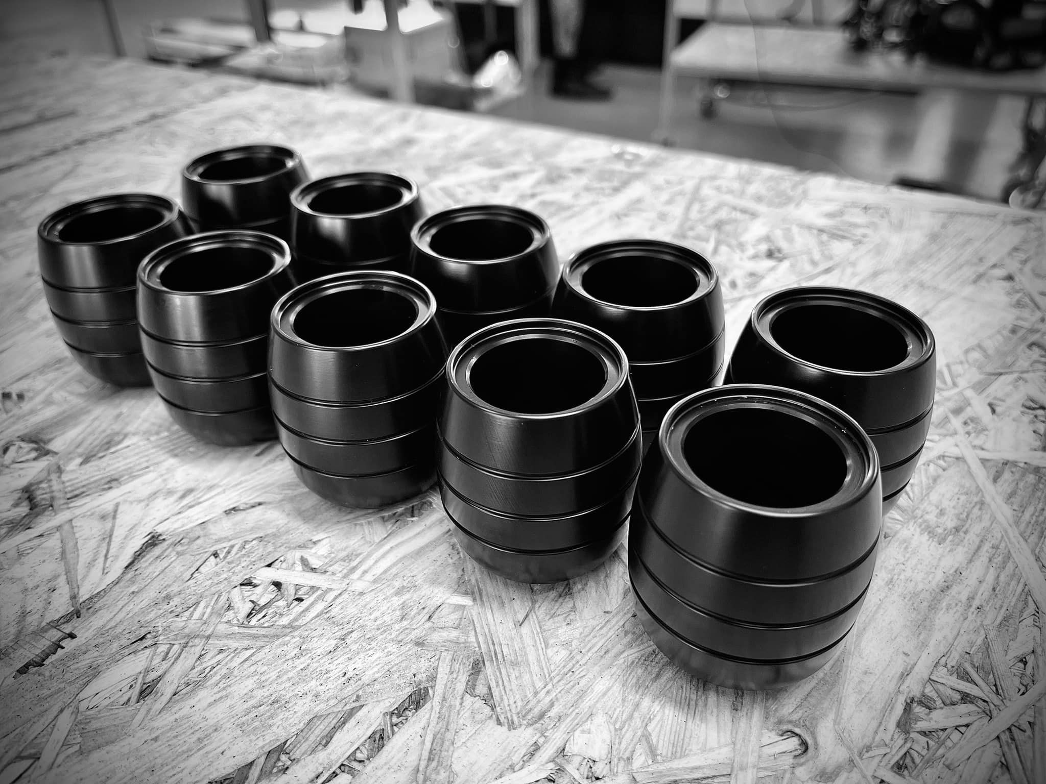 Czarno-białe zdjęcie dziesięciu cylindrycznych, metalowych elementów z rowkami, prawdopodobnie tulejek lub pierścieni, ułożonych na płycie OSB, z rozmytym tłem warsztatowym.