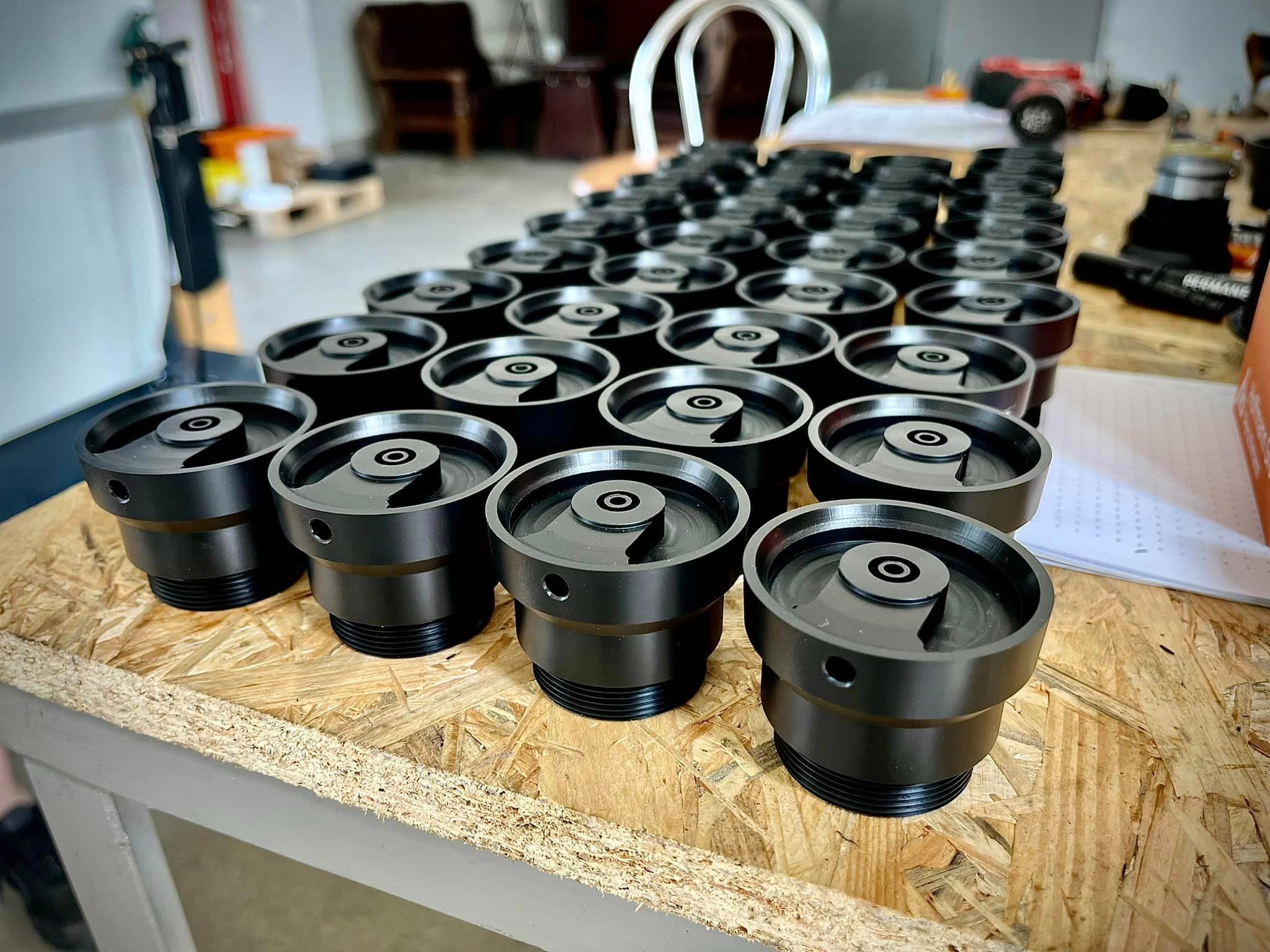 Rząd precyzyjnie wykonanych, czarnych, metalowych elementów z gwintem, ułożonych na płycie OSB. Widoczne otwory boczne i centralne wgłębienie w każdym elemencie. Tło rozmyte, ukazujące inne...