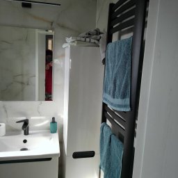 Rafał Maiwald - Nowoczesna łazienka z umywalką nablatową, czarną armaturą, białymi szafkami i czarnym grzejnikiem drabinkowym z powieszonymi turkusowymi ręcznikami; odbicie osoby robiącej zdjęcie w lustrze.