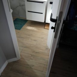 Rafał Maiwald - Otwarta biała łazienkowa z czarną armaturą, umywalką z szafką, marmurowymi ścianami i drewnopodobną podłogą z płytek, widziana zza uchylonych drzwi.