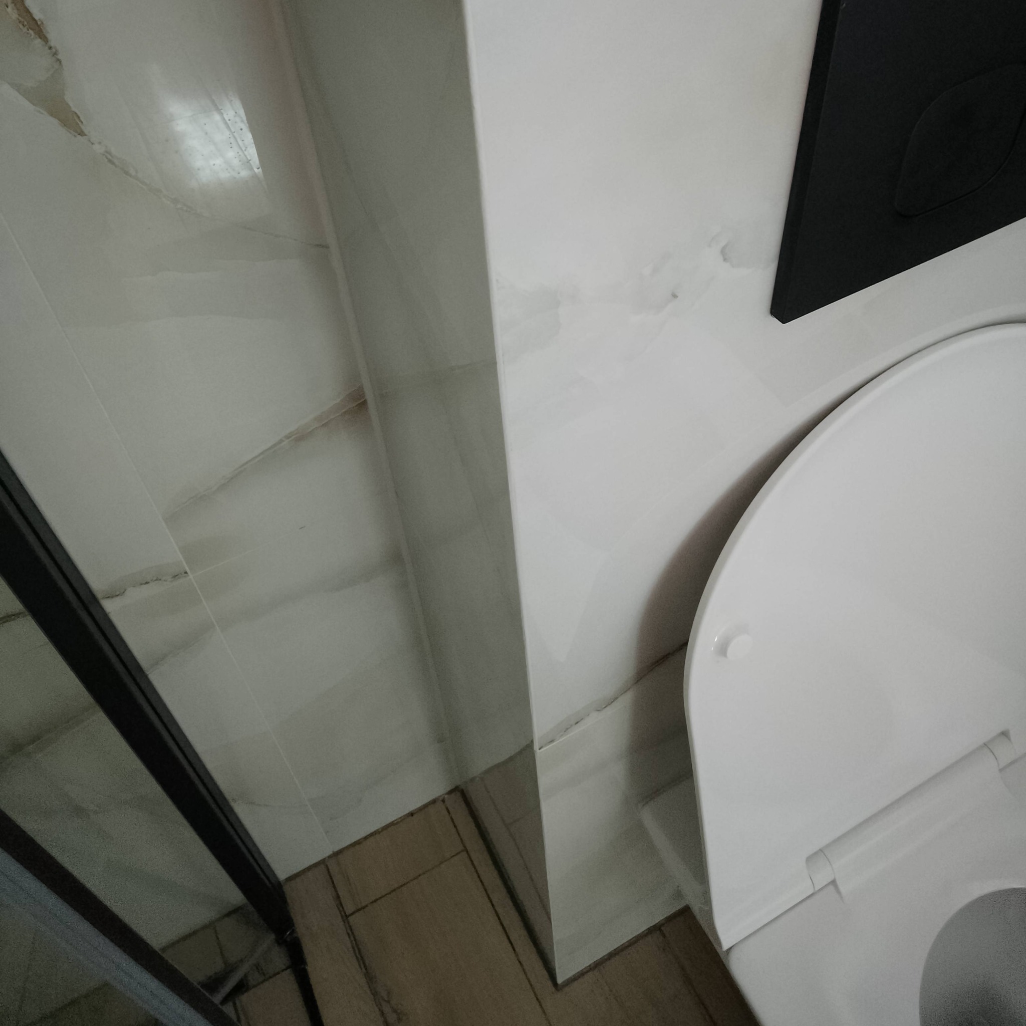 Fragment wykończonej łazienki z widoczną toaletą, płytkami imitującymi marmur na ścianach i drewnopodobną podłogą, czarny przycisk spłuczki, ujęcie z góry.