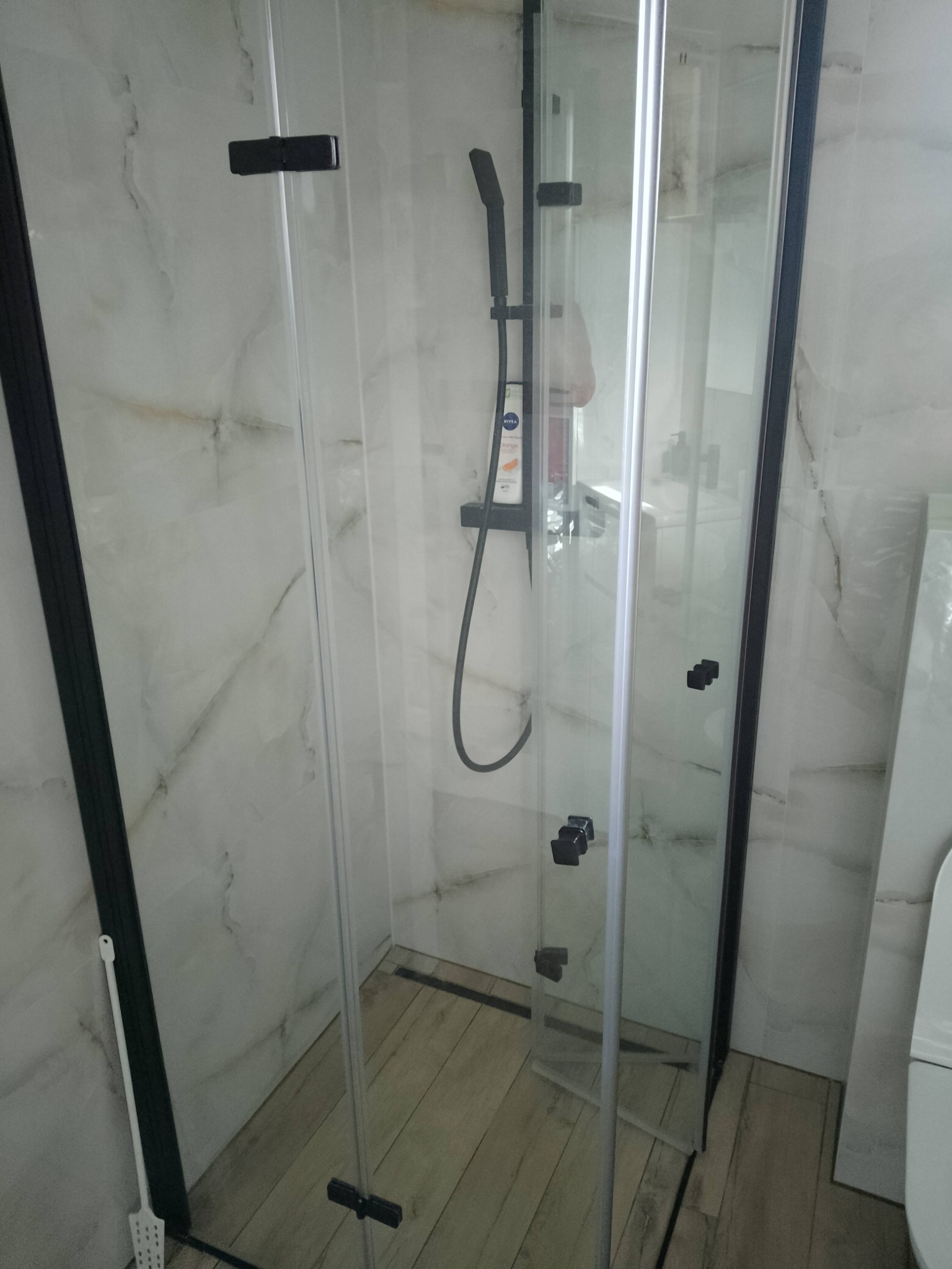 Nowoczesna kabina prysznicowa z czarnymi profilami i armaturą, jasne płytki imitujące marmur na ścianach, drewnopodobna podłoga, widoczna chemia gospodarcza na półce w kabinie.