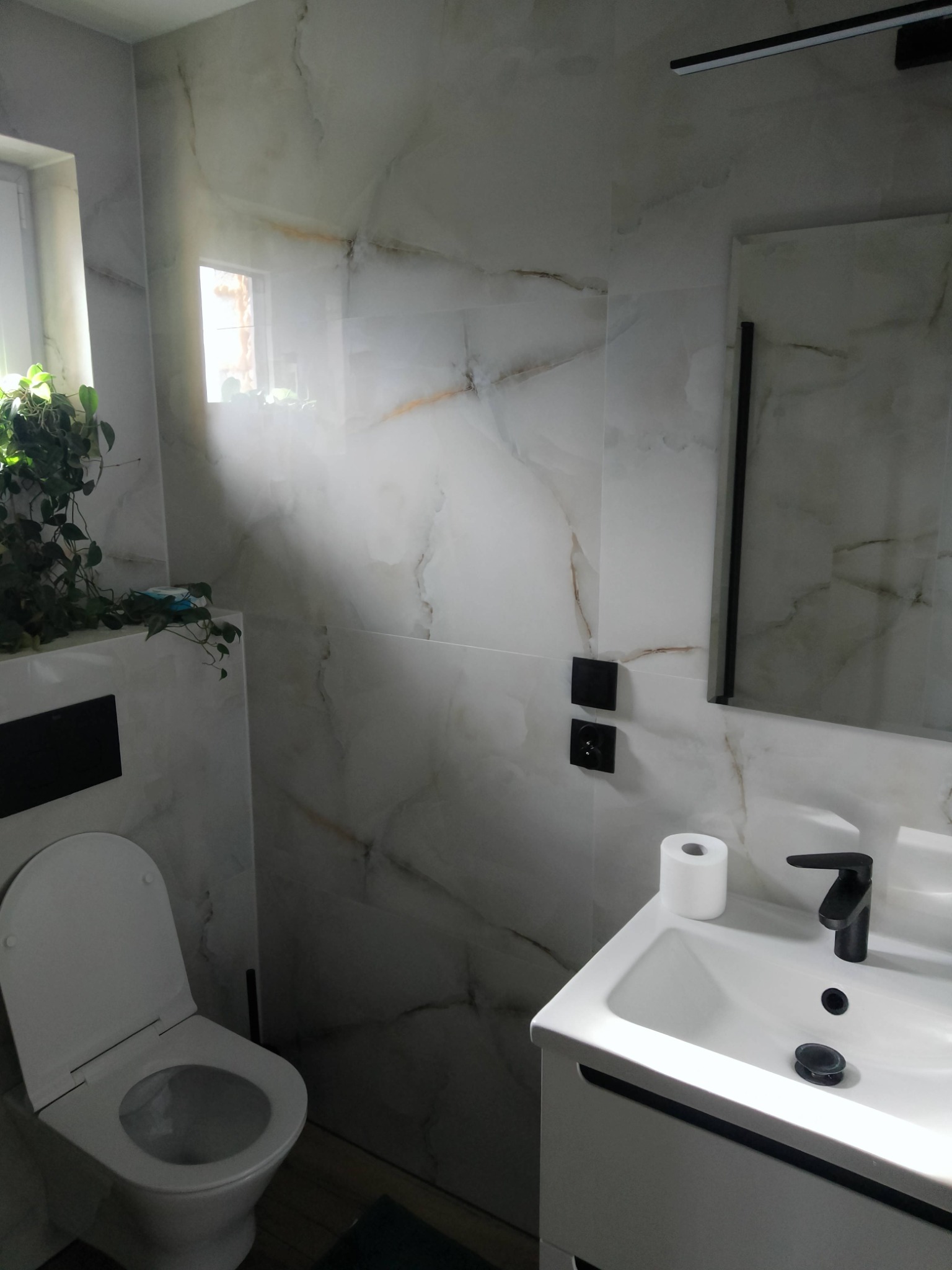 Nowoczesna łazienka z marmurowymi płytkami, wiszącą toaletą i umywalką z czarną armaturą, oświetlona światłem dziennym z okna z zielonymi roślinami.