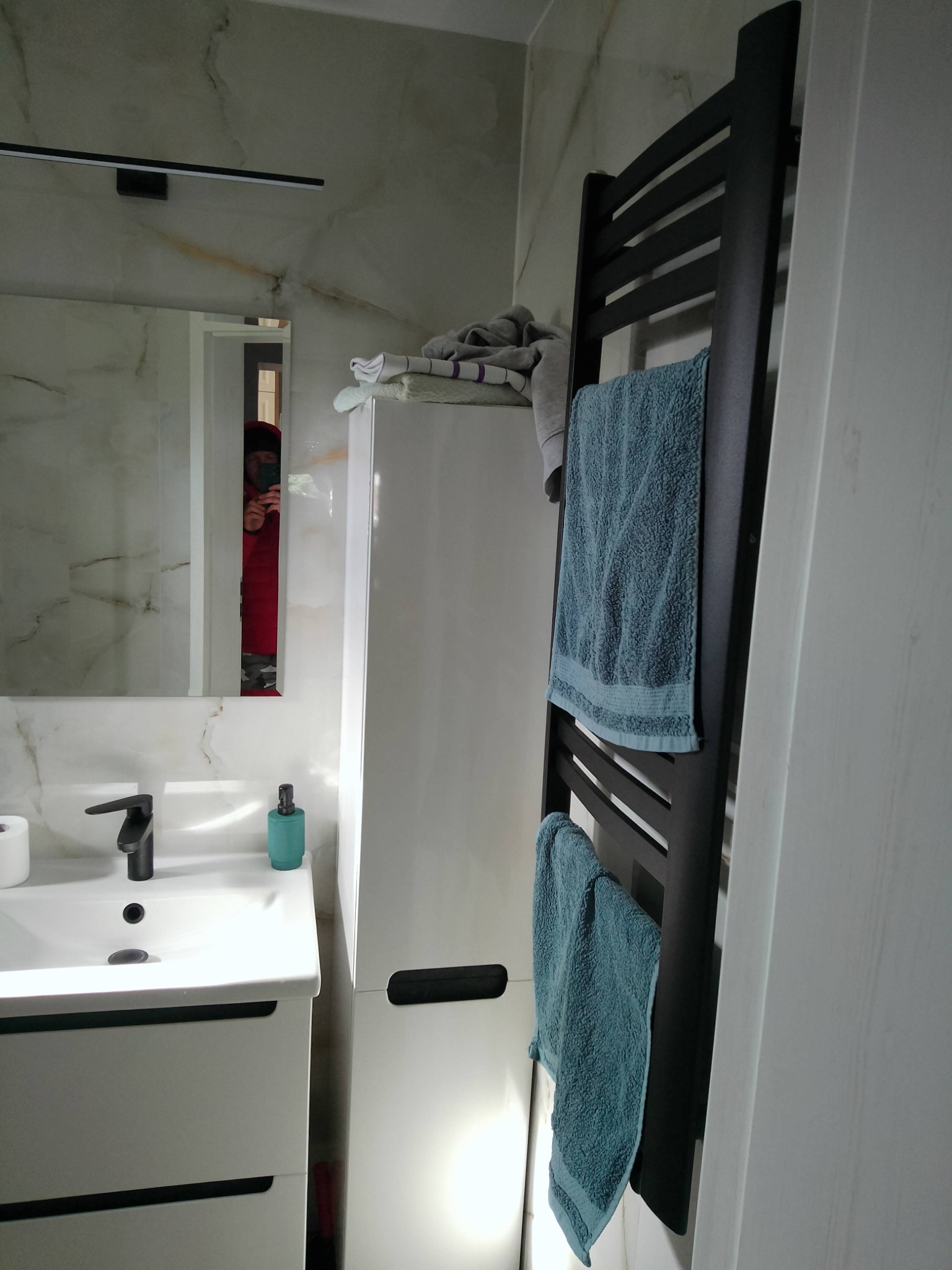 Nowoczesna łazienka z umywalką nablatową, czarną armaturą, białymi szafkami i czarnym grzejnikiem drabinkowym z powieszonymi turkusowymi ręcznikami; odbicie osoby robiącej zdjęcie w lustrze.