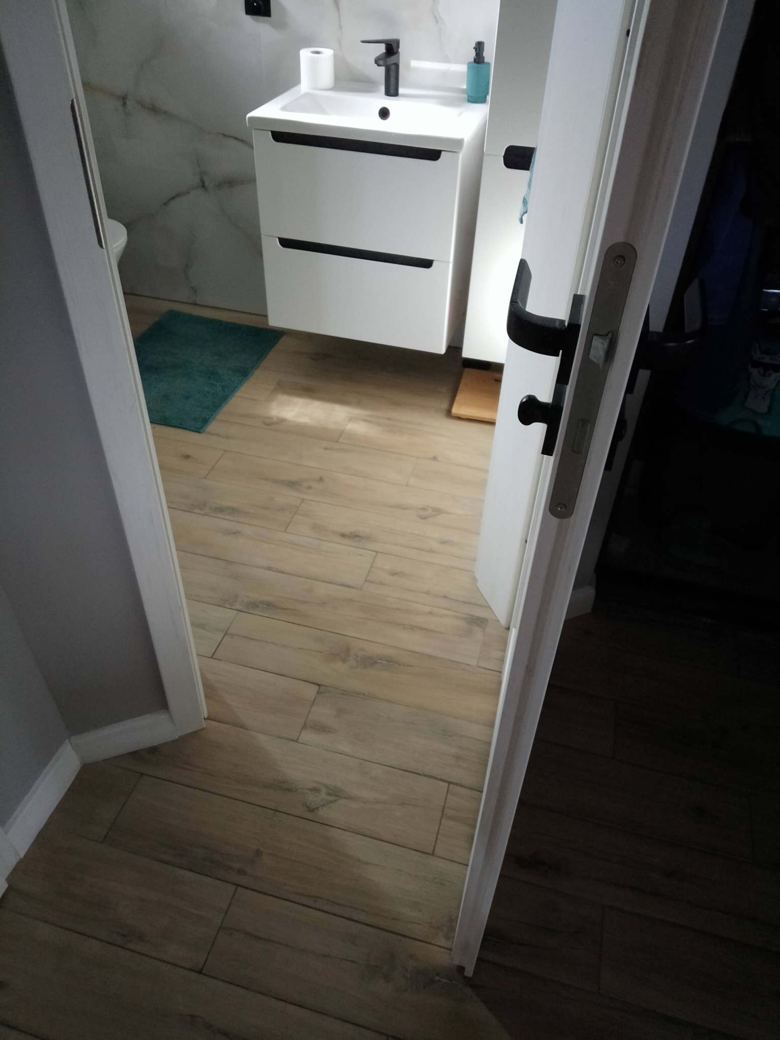 Otwarta biała łazienkowa z czarną armaturą, umywalką z szafką, marmurowymi ścianami i drewnopodobną podłogą z płytek, widziana zza uchylonych drzwi.