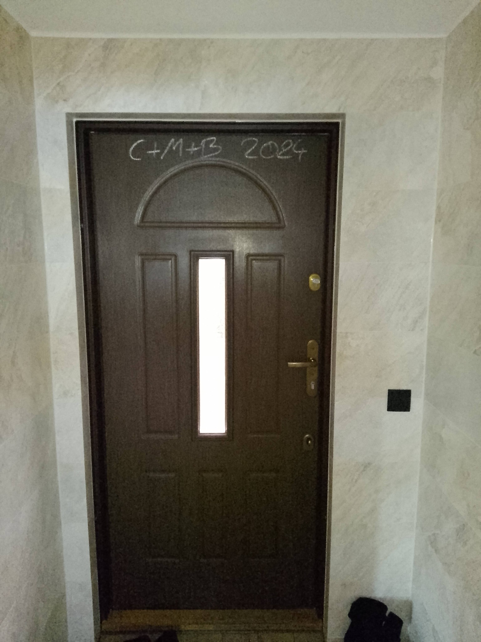 Ciemnobrązowe drzwi wejściowe z szybą, otoczone beżowymi płytkami imitującymi marmur, z napisem 'C+M+B 2024' wykonanym kredą nad górną częścią drzwi.