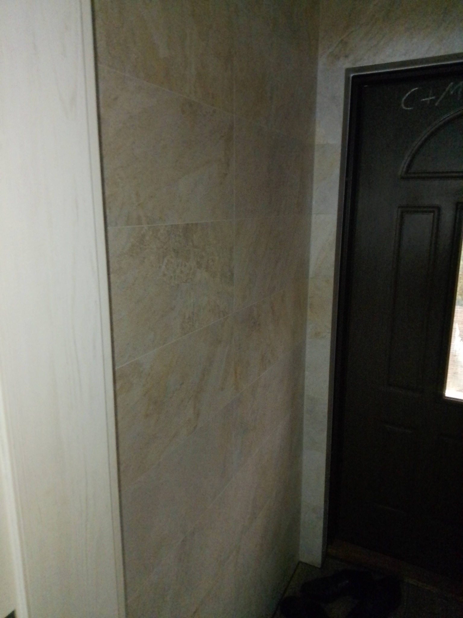 Fragment łazienki z beżowymi płytkami imitującymi kamień na ścianie, widoczna czarna ościeżnica drzwi z napisem 'C+M' oraz fragment białej szafki.