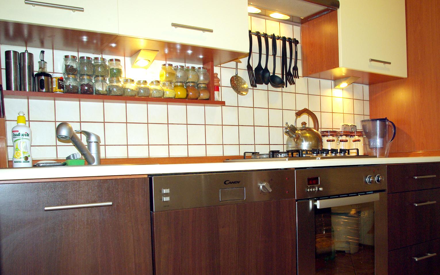 Kuchnia z drewnianymi szafkami, białymi górnymi szafkami, ceramiczną płytą grzewczą, czajnikiem na palniku, półką z przyprawami w słoikach i oświetleniem punktowym pod szafkami.